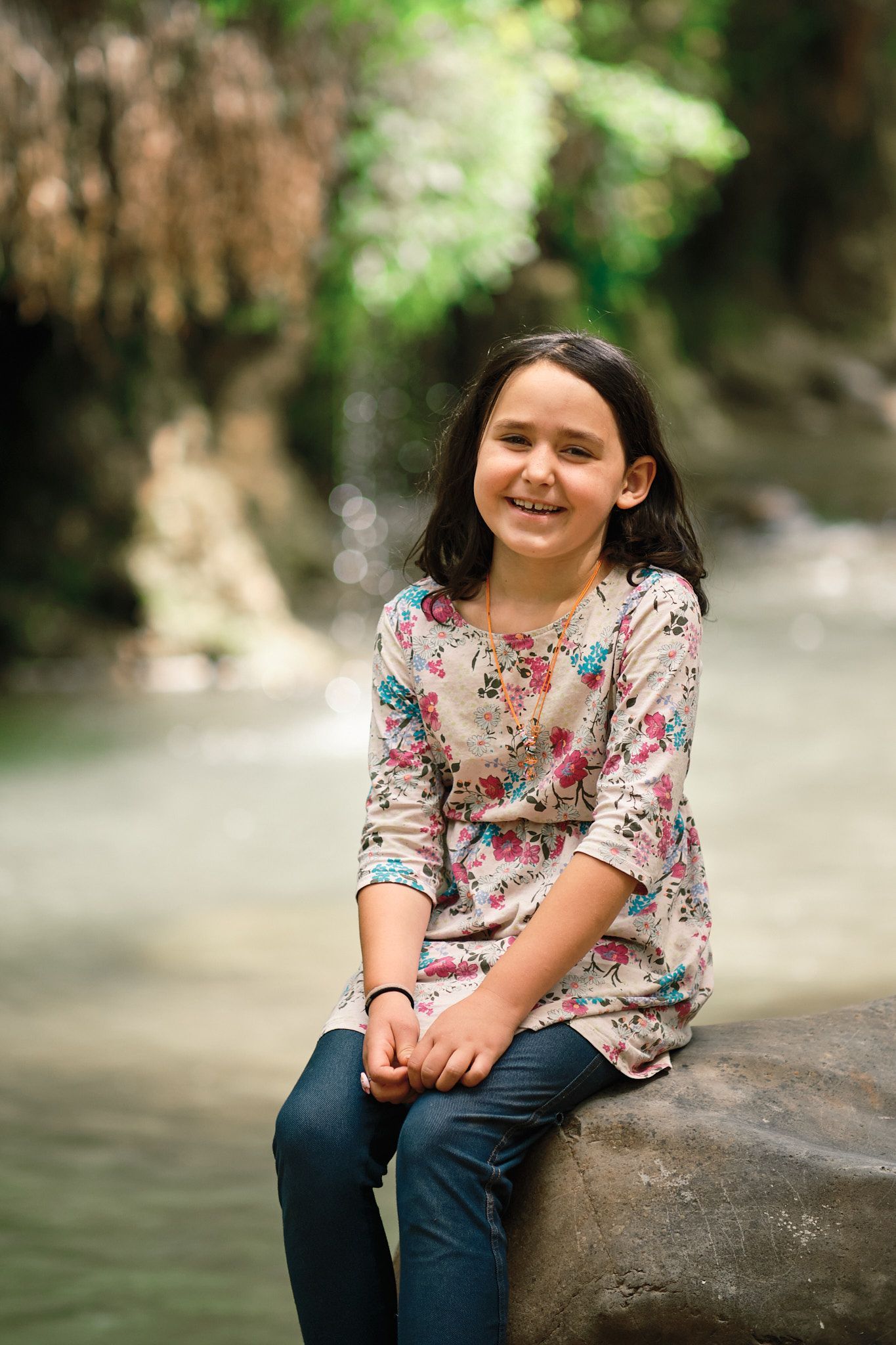 A smiling person wearing a floral top and dark pants sitting on a rock by a stream in a forest.