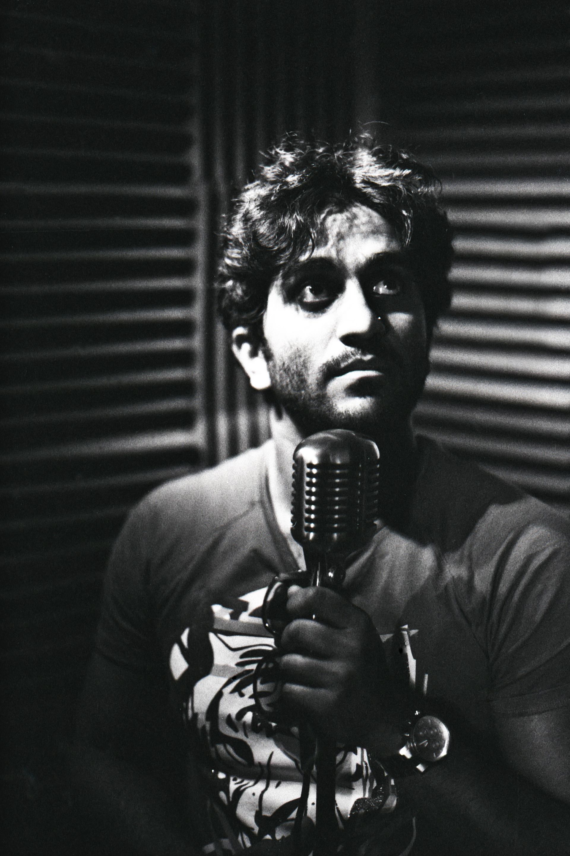 A person stands in a studio, looking upward while holding a vintage-style microphone in front of them. Black and white.