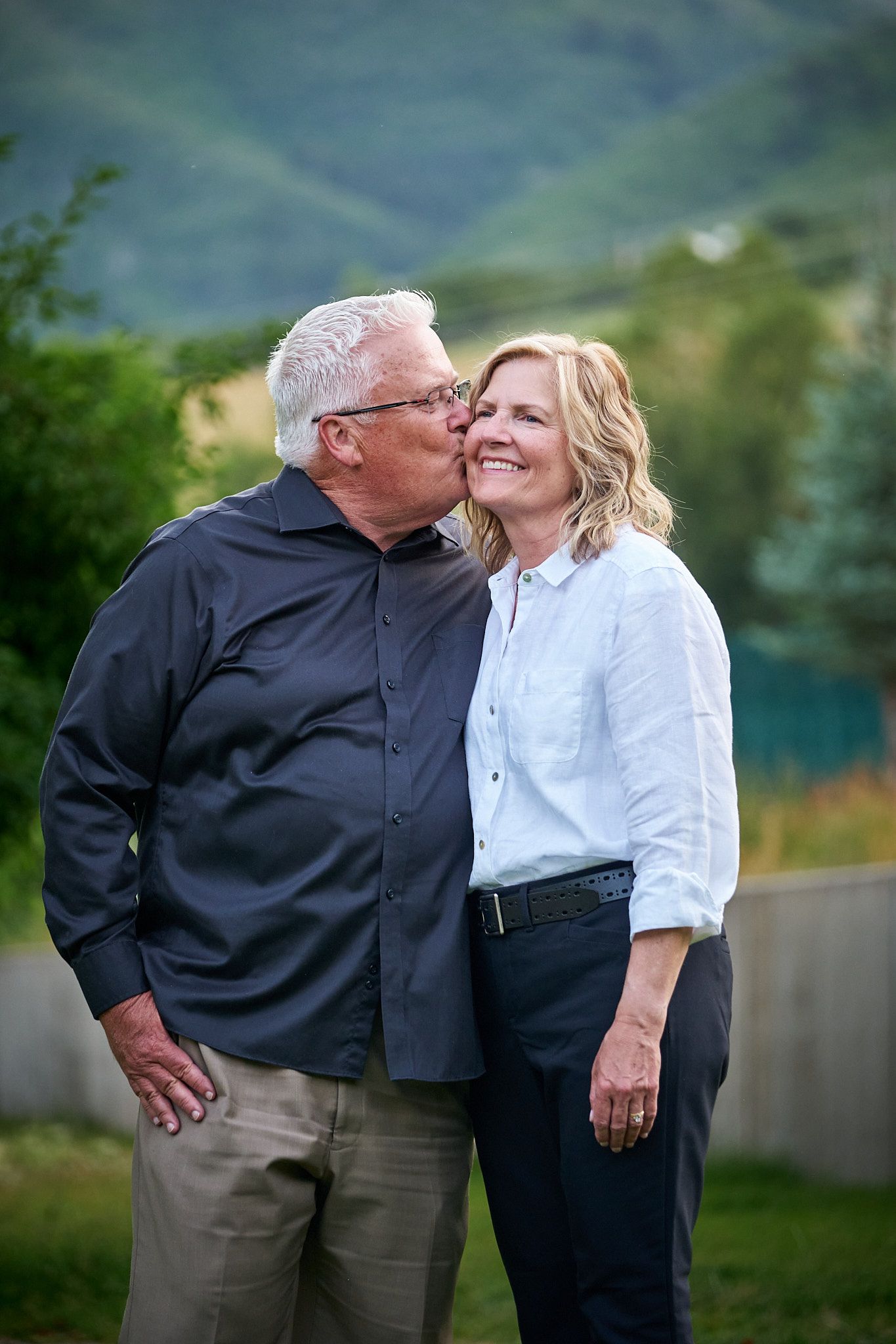 An older person in a black shirt kisses the cheek of a smiling person in a white shirt outdoors against a hilly background.