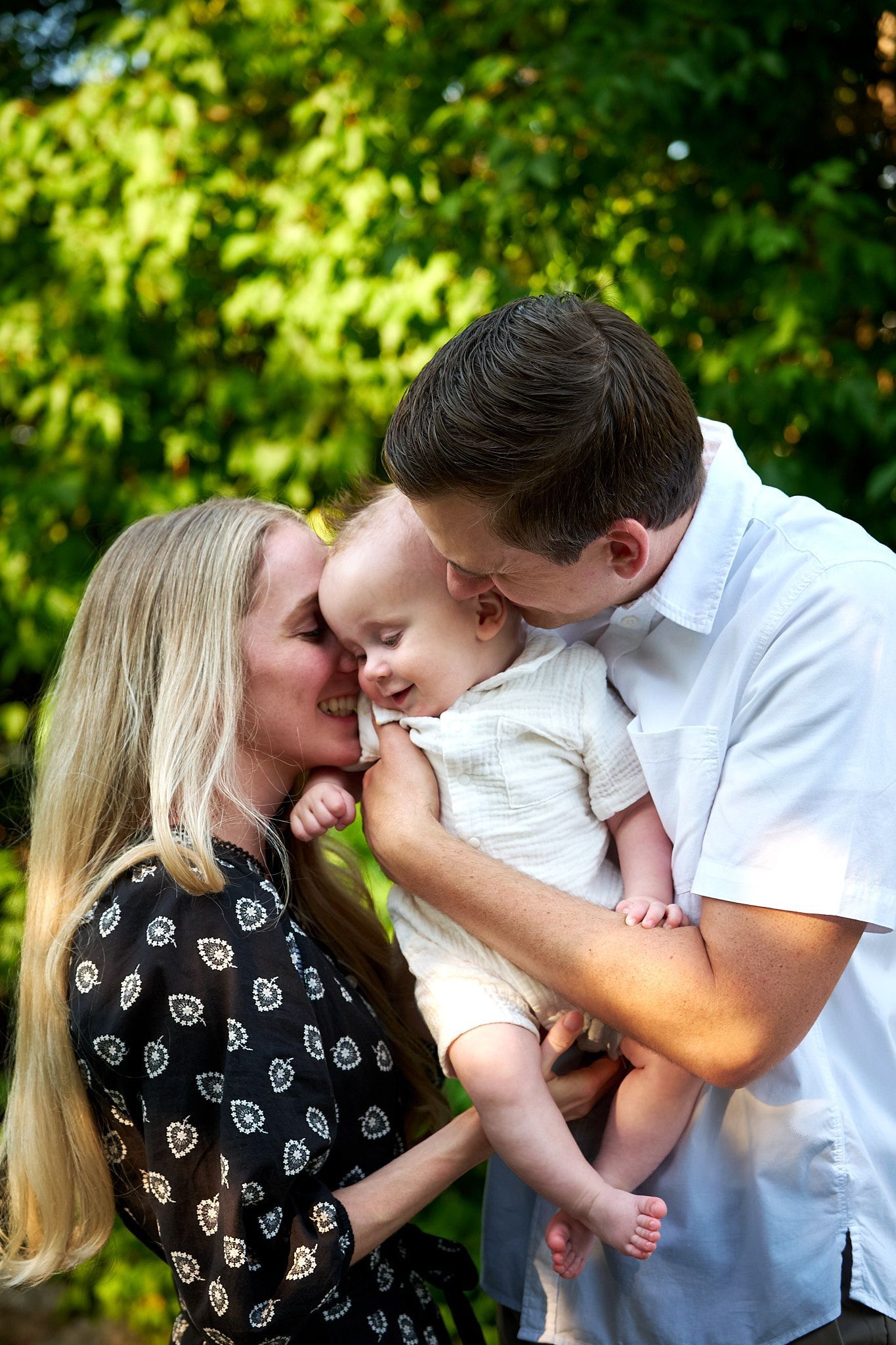 A couple cradles their smiling baby outdoors, surrounded by soft green foliage.
