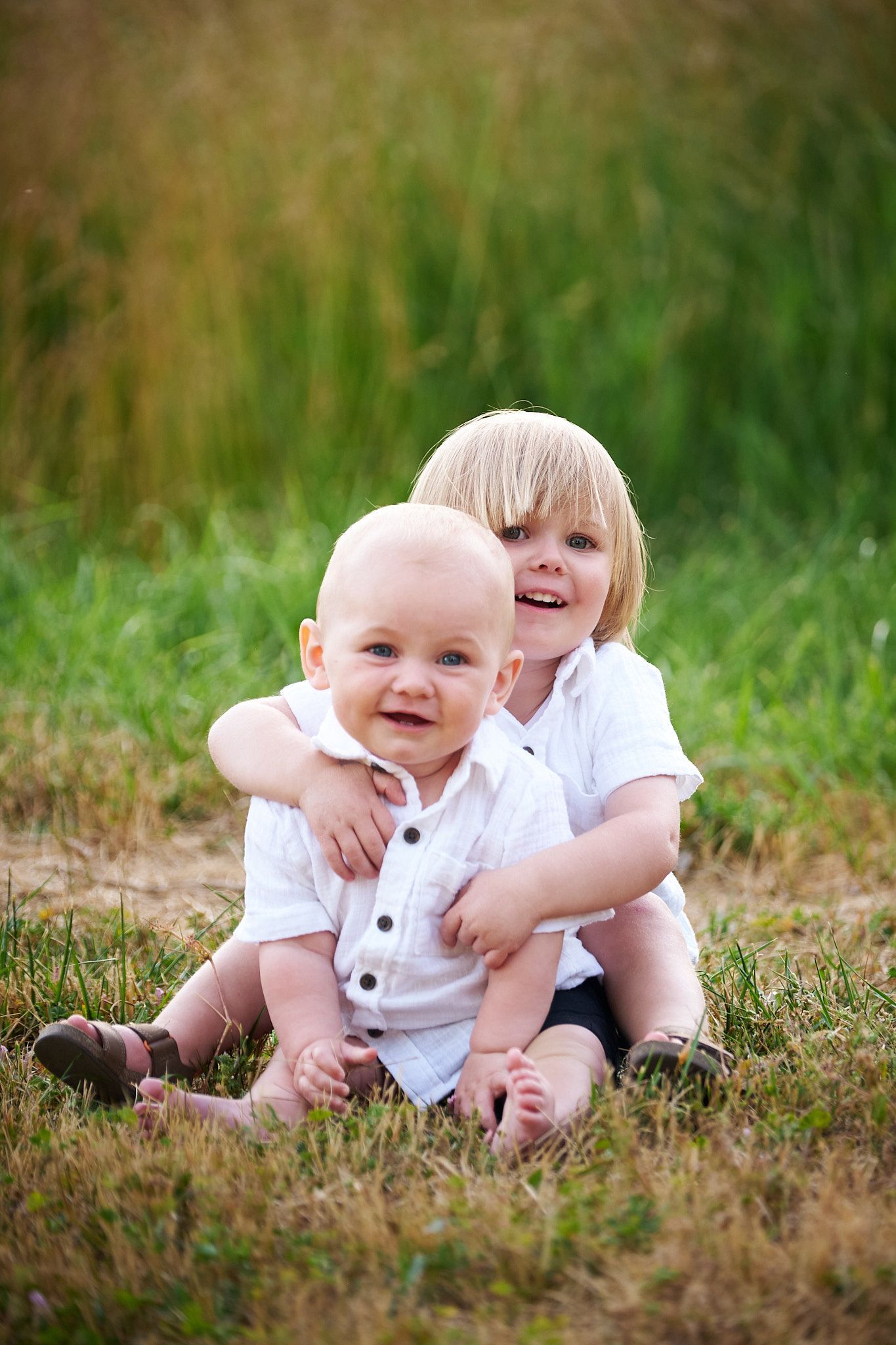 Two children in white shirts sit together in a grassy field, with one hugging the other from behind.