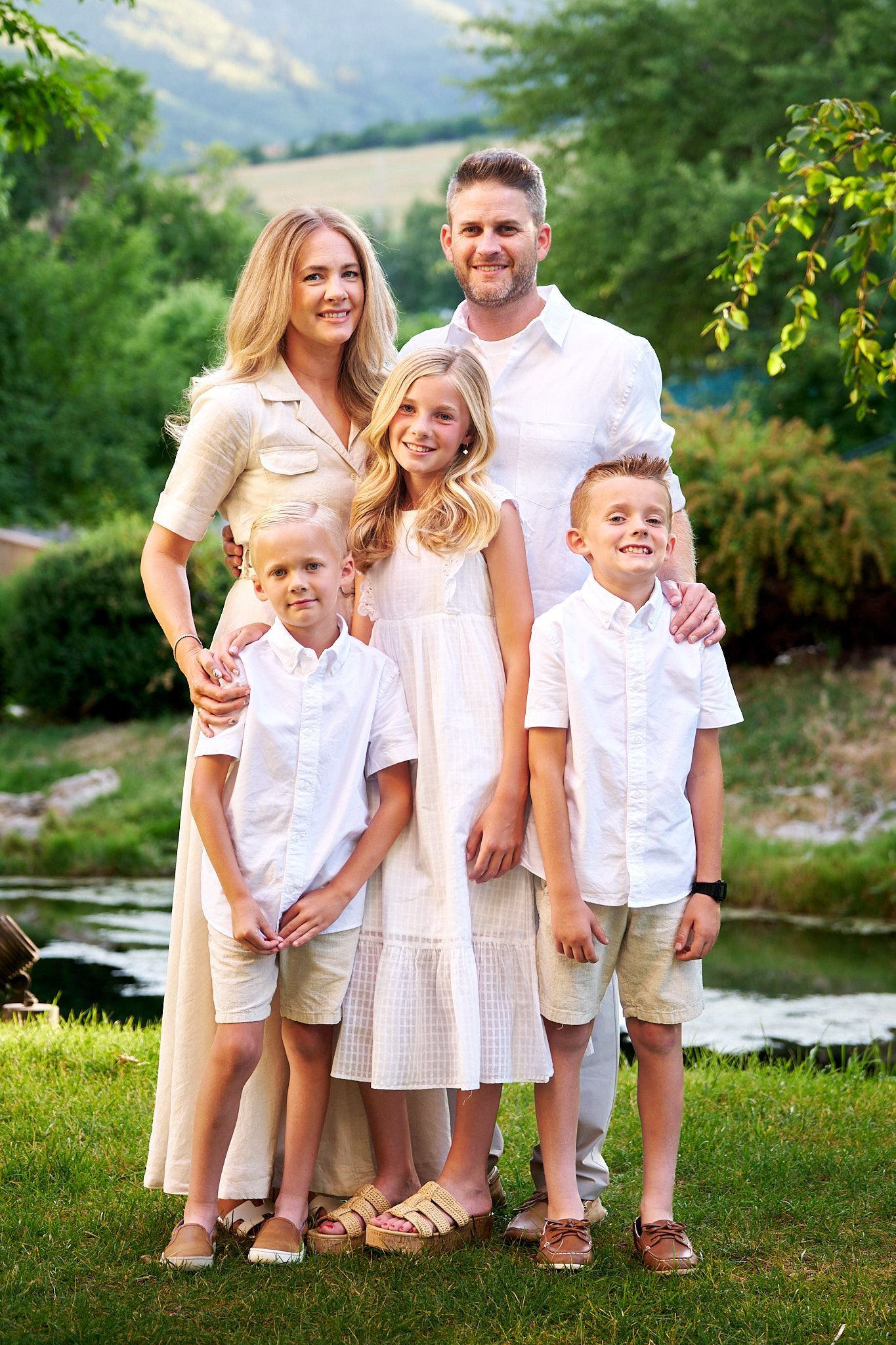 A family stands together in an outdoor setting, dressed in white, positioned in front of greenery and a small pond.