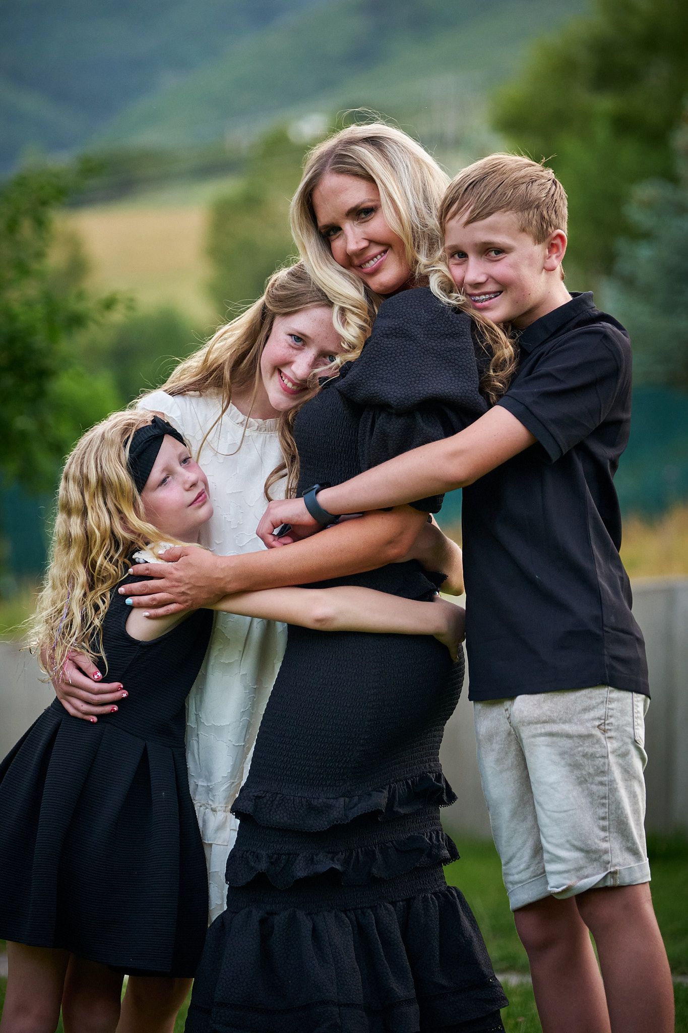 A woman stands outdoors, smiling as she is embraced by three young people, all dressed in formal black and white attire.