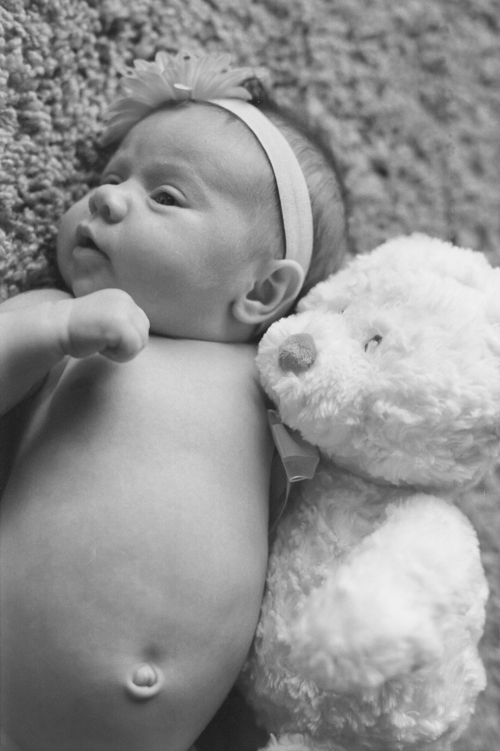A baby wearing a headband lies on a textured surface next to a white teddy bear.