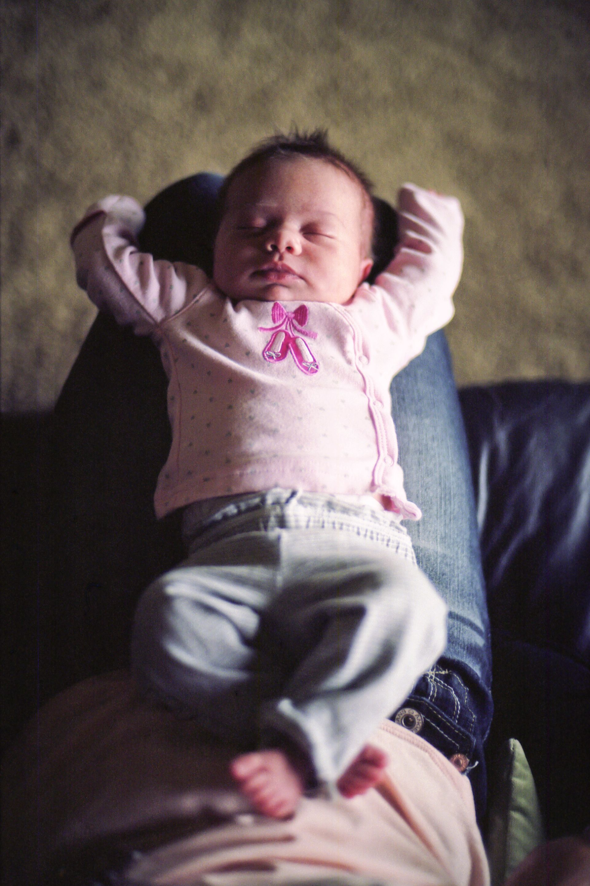 An infant wearing a pink long-sleeved shirt and gray pants sleeping while lying on someone's lap.