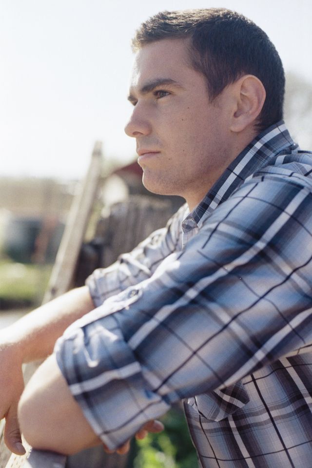 A person wearing a blue and white plaid shirt leans against a fence, looking into the distance with a thoughtful gaze.