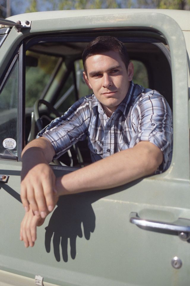 A person in a blue plaid shirt resting their arms on the open window of a light green truck, looking at the camera.