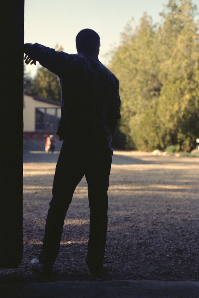 A person in silhouette stands in a doorway, leaning against the frame while looking out at a gravel yard and trees.