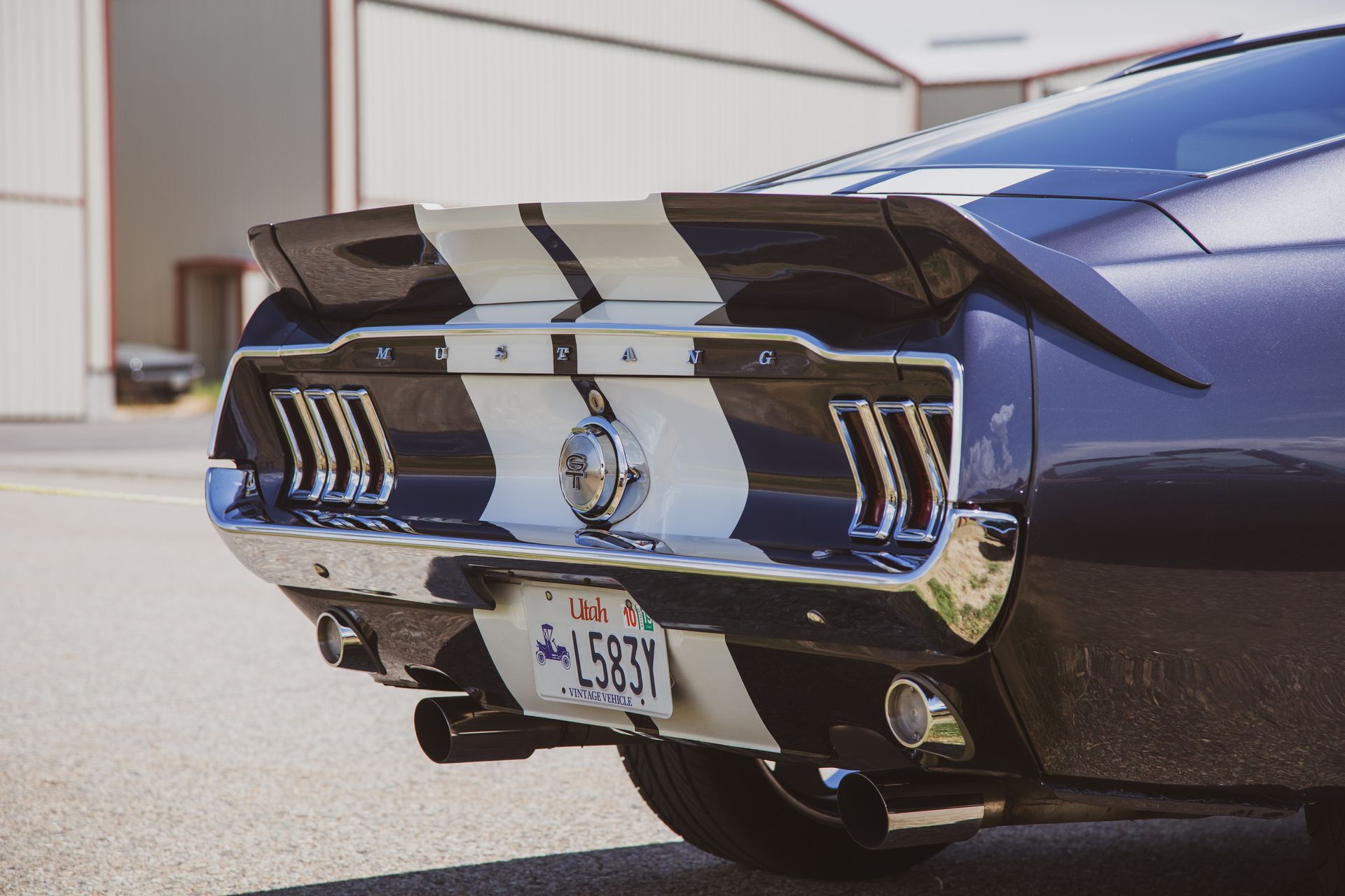 Rear view of a dark blue classic Ford Mustang with white racing stripes and a license plate, parked outdoors.