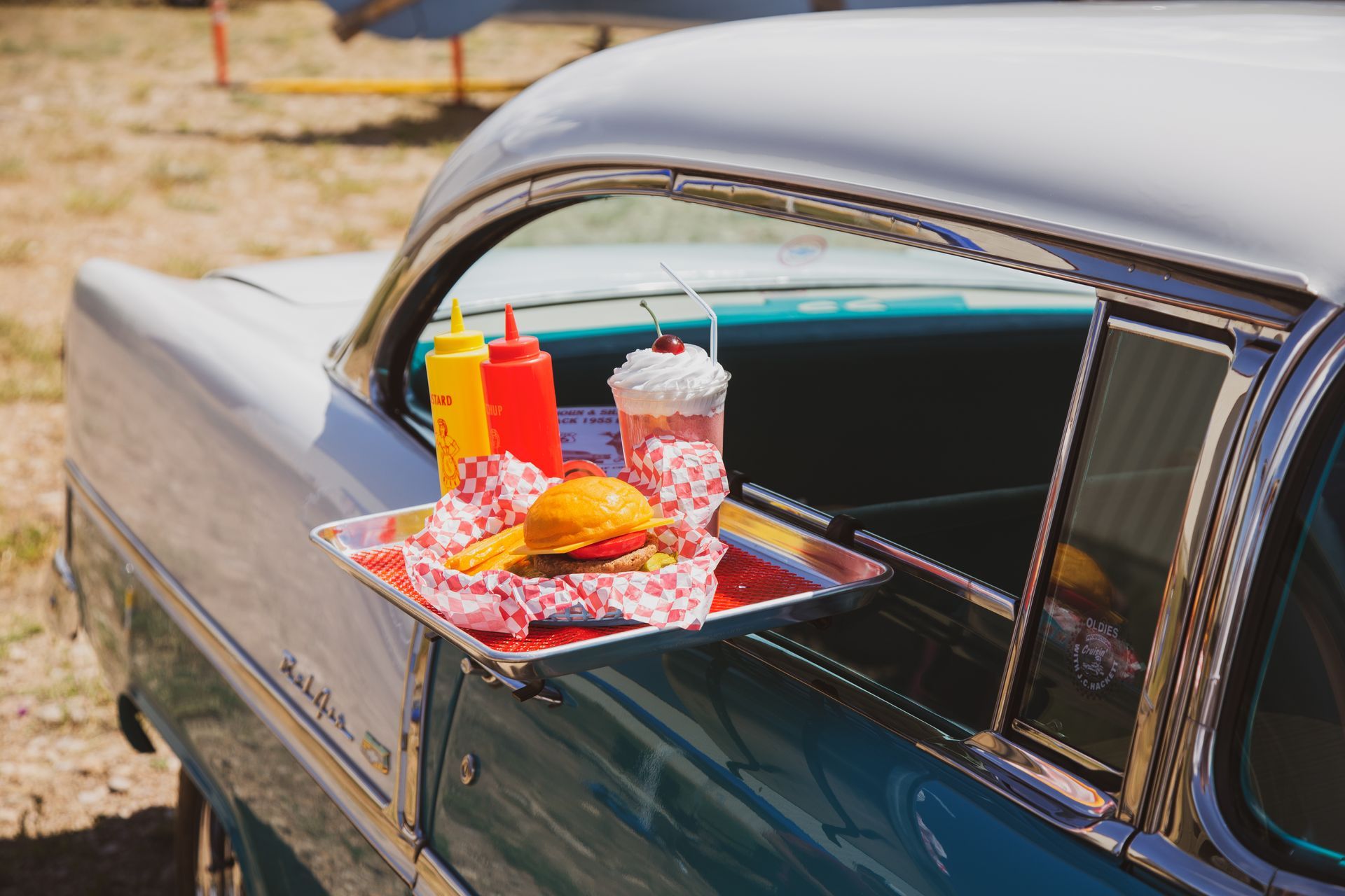 A tray with a hamburger, fries, condiments, and a milkshake sits on the window of a teal and white vintage car.