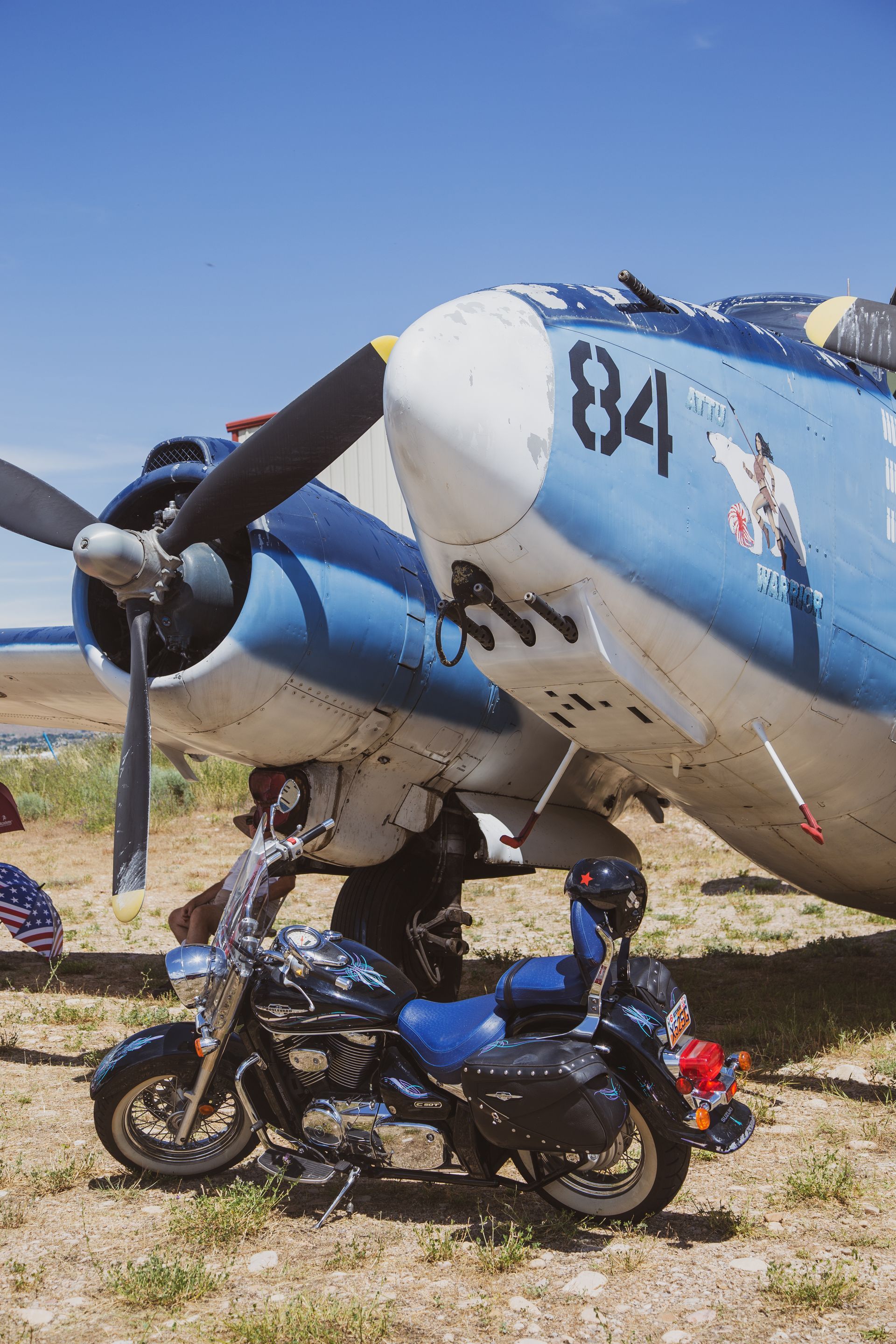 A black motorcycle parked on dry ground beneath the nose and propeller of a vintage blue and white military aircraft.