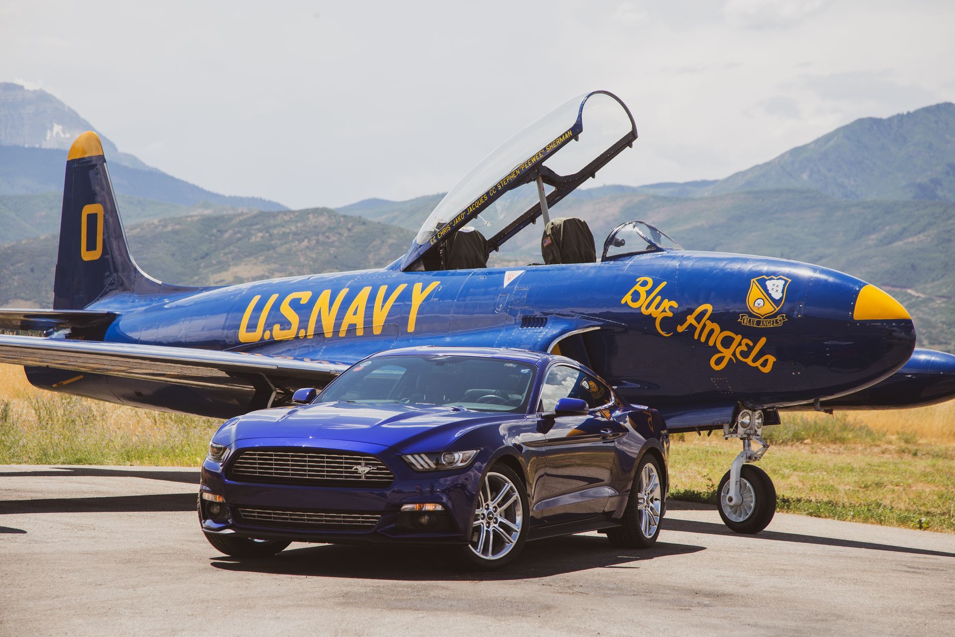 A blue Ford Mustang parked in front of a blue U.S. Navy Blue Angels jet on a paved tarmac with mountains in the background.