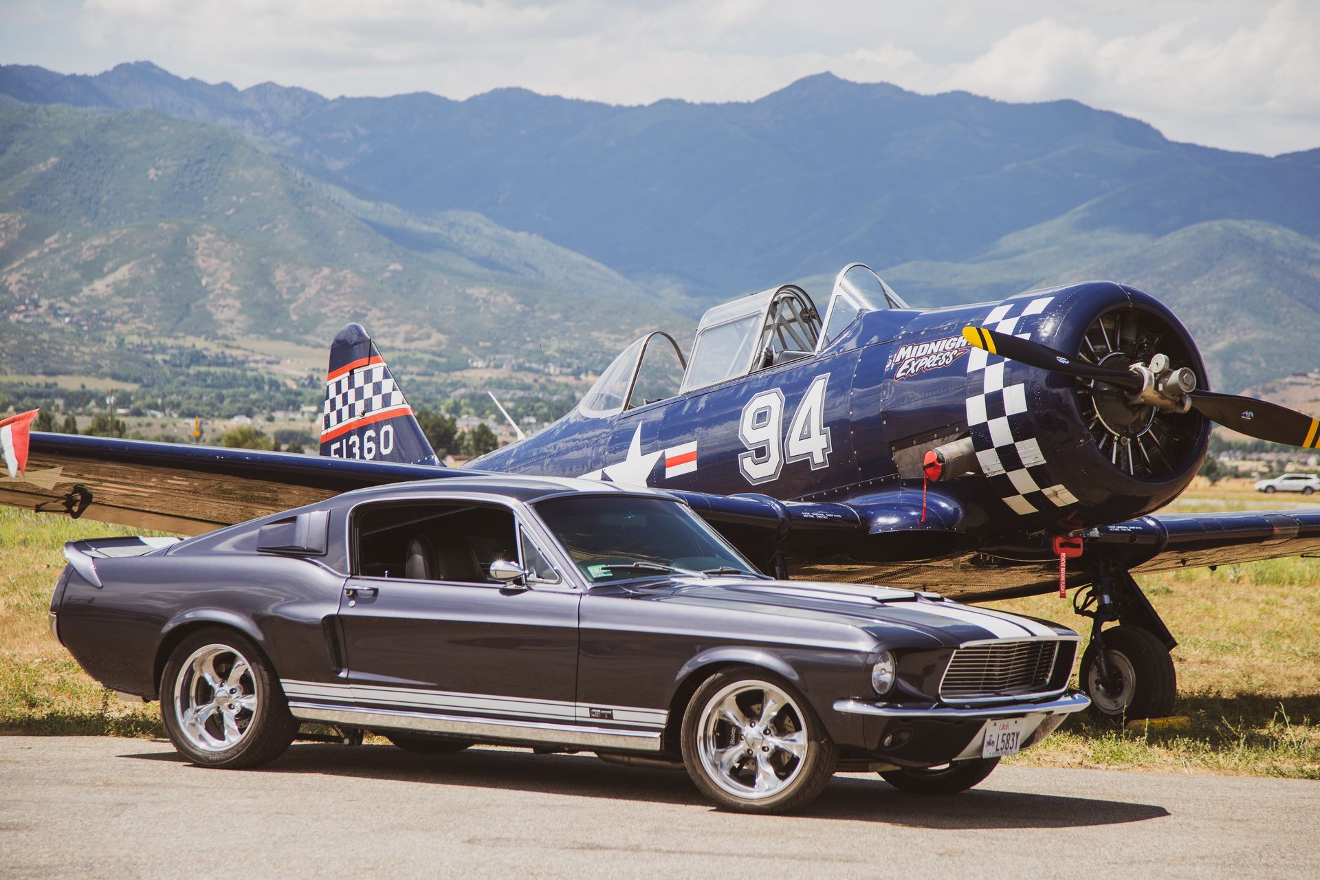 A dark gray Ford Mustang parked in front of a blue vintage aircraft with 
