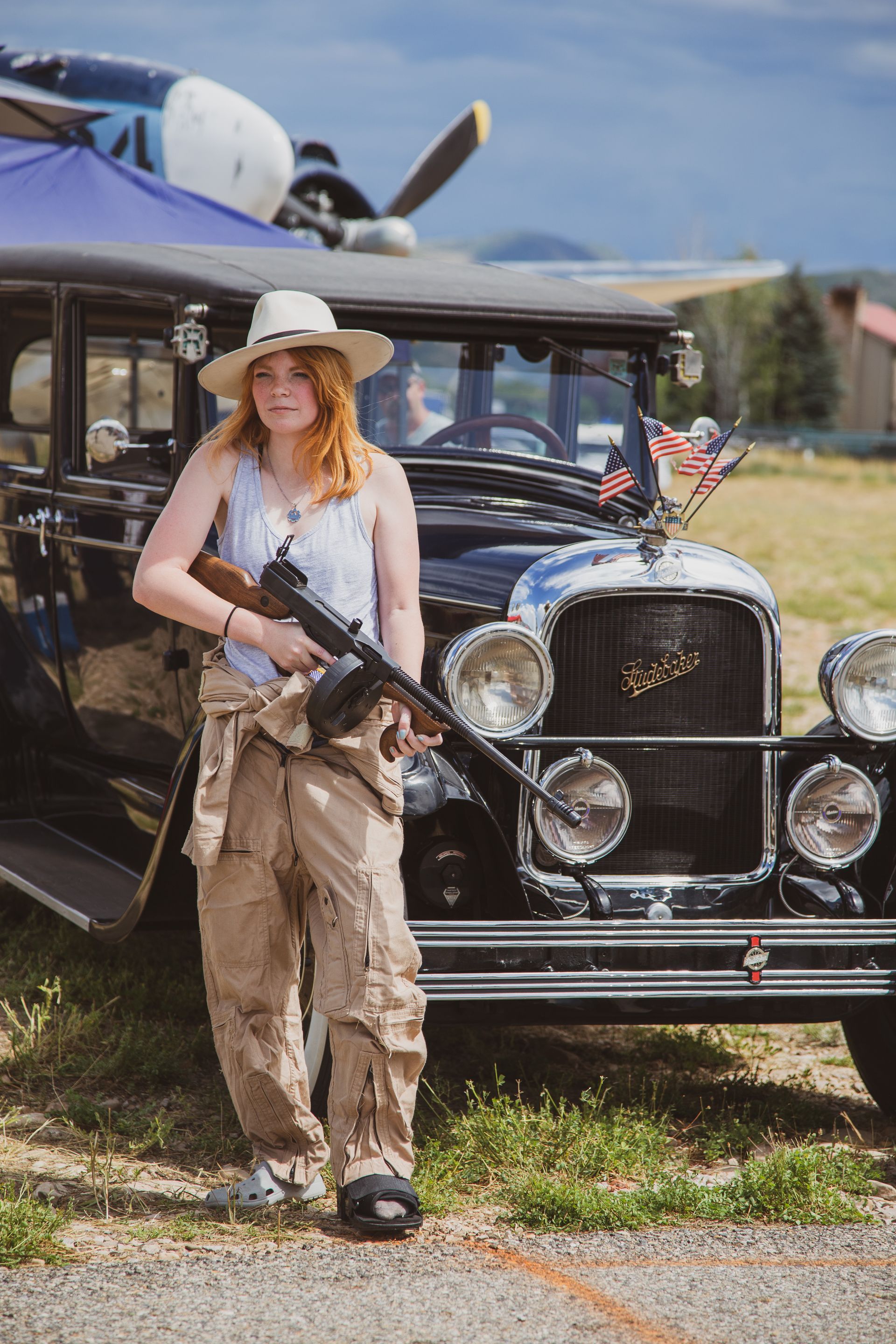 A person wearing a hat and tan overalls holds a prop machine gun in front of a vintage car parked near an airplane.