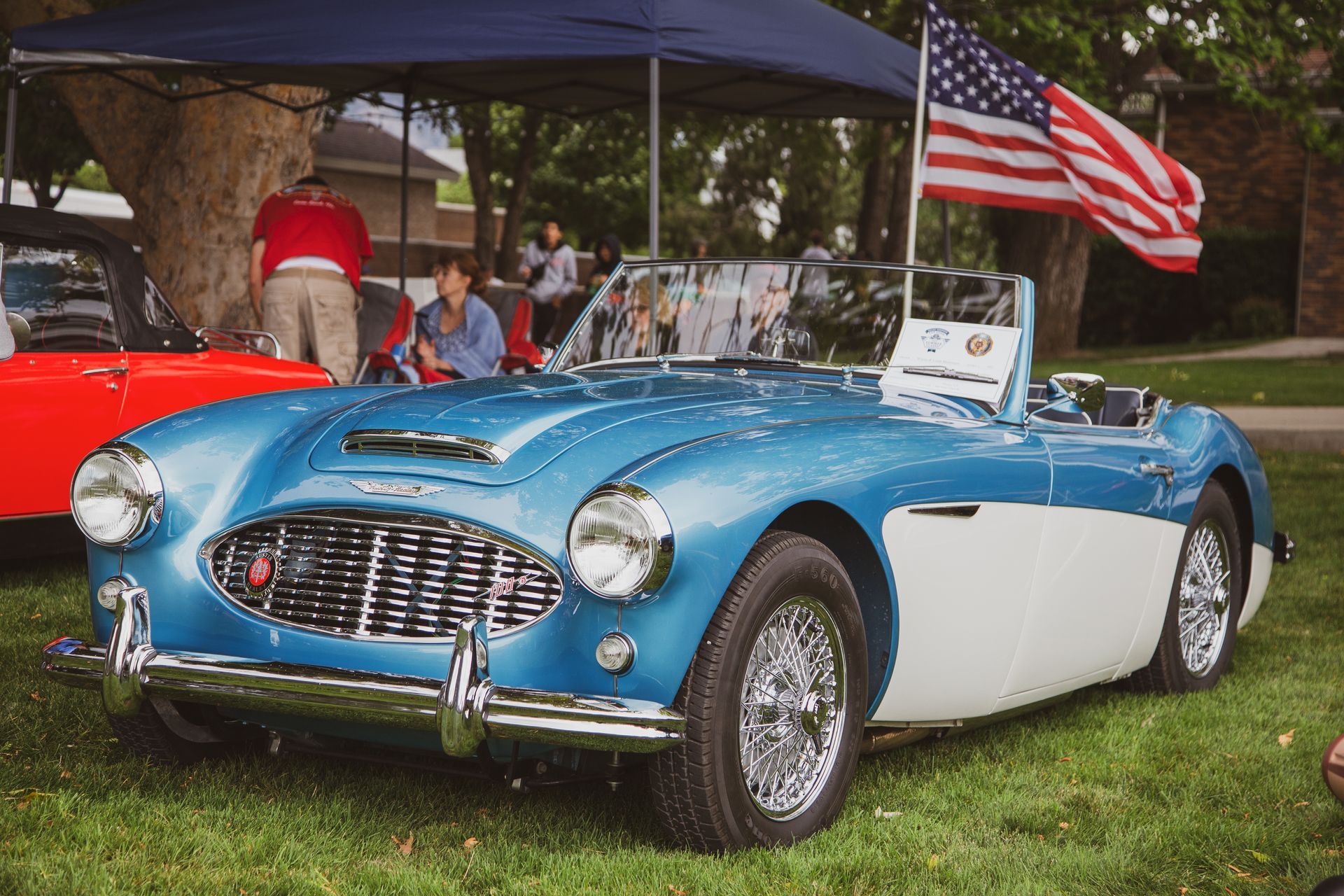 A blue and white Austin-Healey vintage sports car parked on grass with an American flag flying behind it.