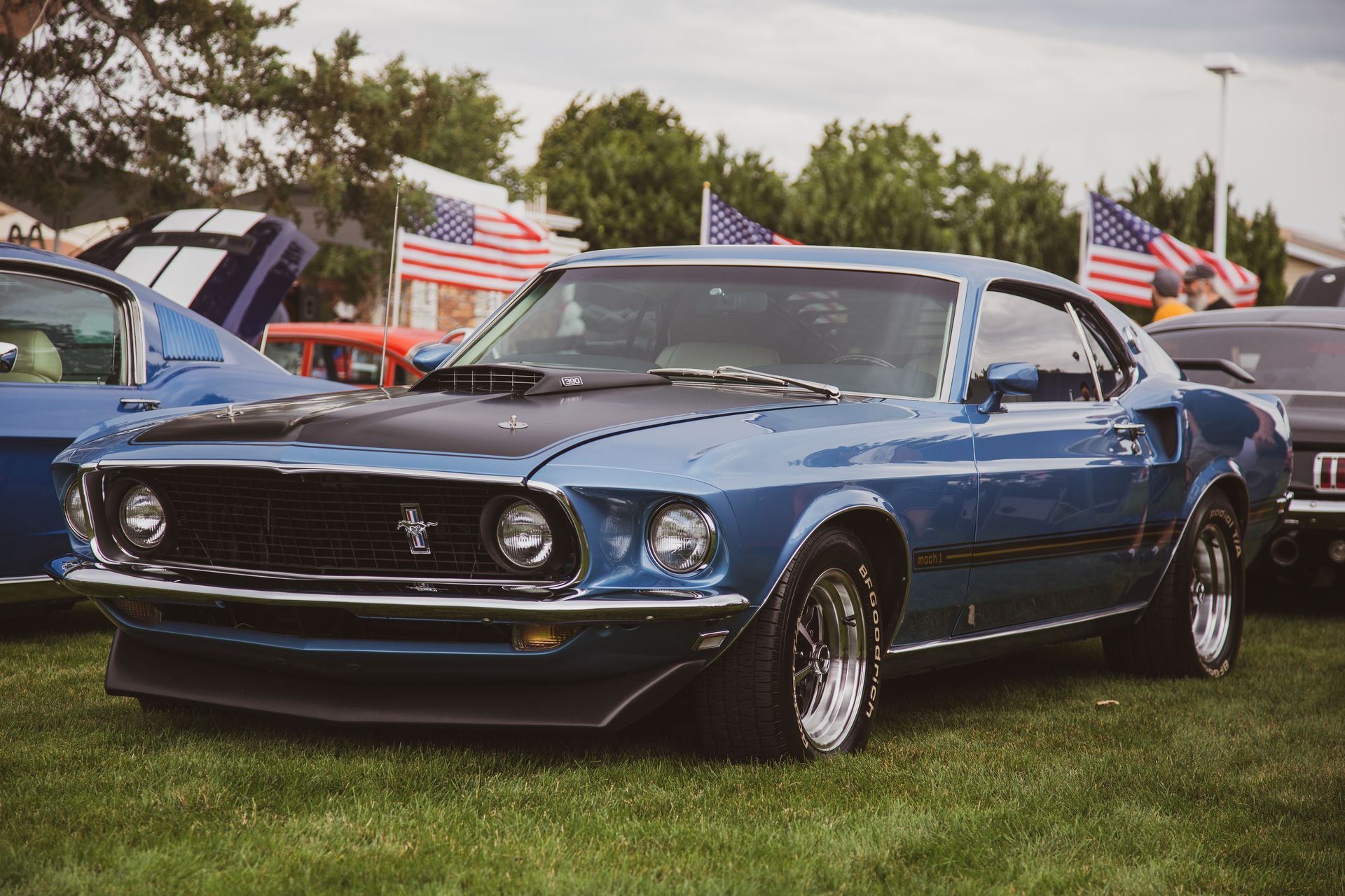 A blue classic Ford Mustang with a black hood at an outdoor car show, with American flags in the background.