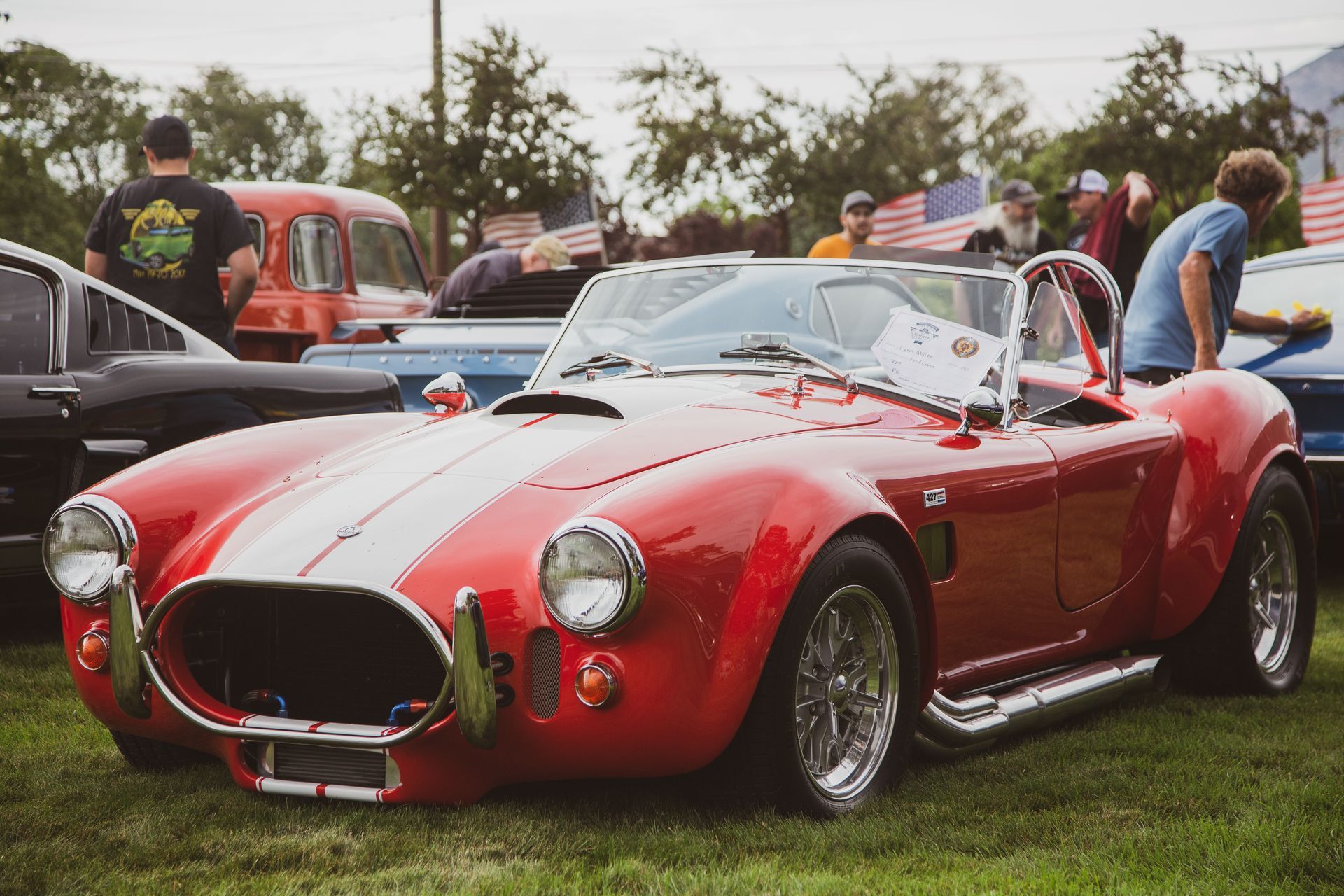 A red classic Shelby Cobra sports car with white racing stripes parked on grass at an outdoor car show.