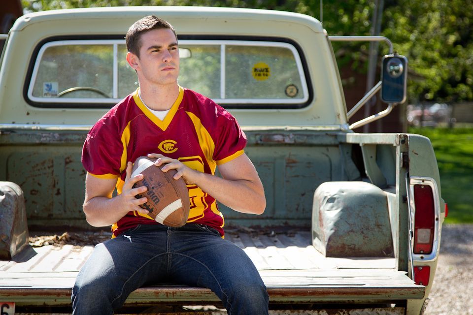 A person wearing a maroon and gold jersey holds a football while sitting on the tailgate of an old white pickup truck.