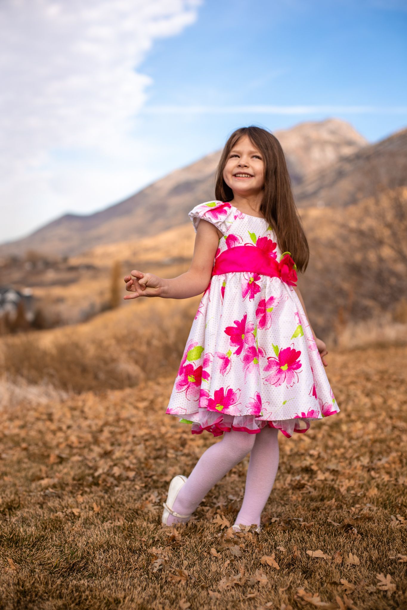 A smiling child in a floral white and pink dress stands on a grassy hillside with mountains in the background.