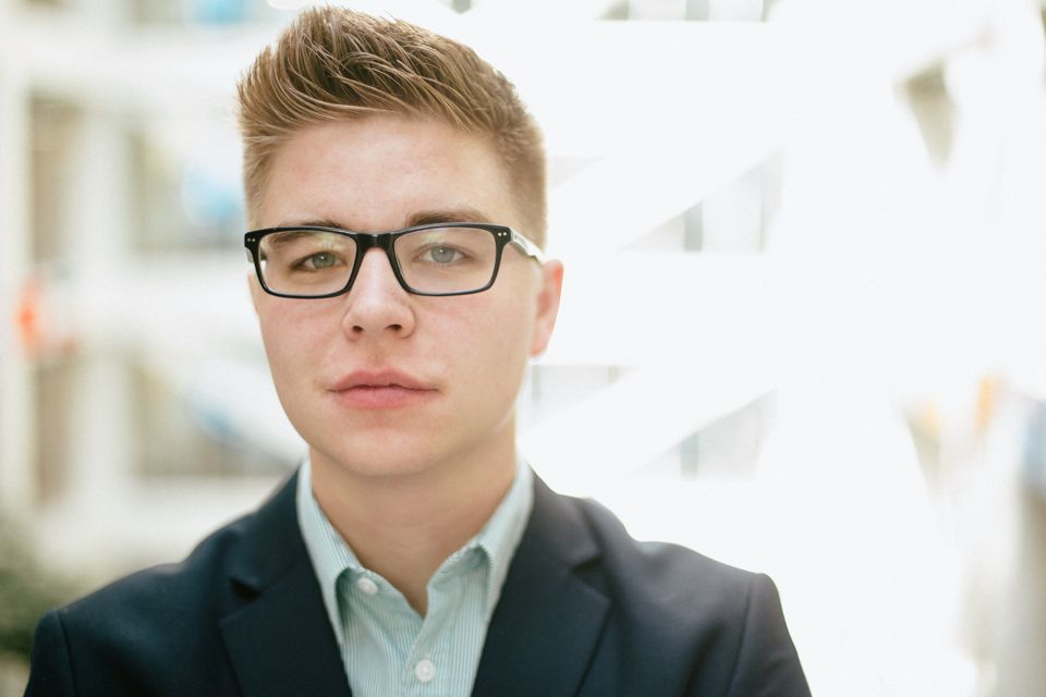 A person with short brown hair and glasses wears a light collared shirt and dark blazer against a bright, blurred backdrop.
