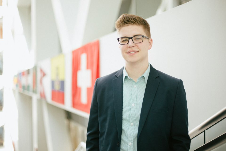 A person in a dark blazer and light shirt stands smiling in front of a wall decorated with several international flags.