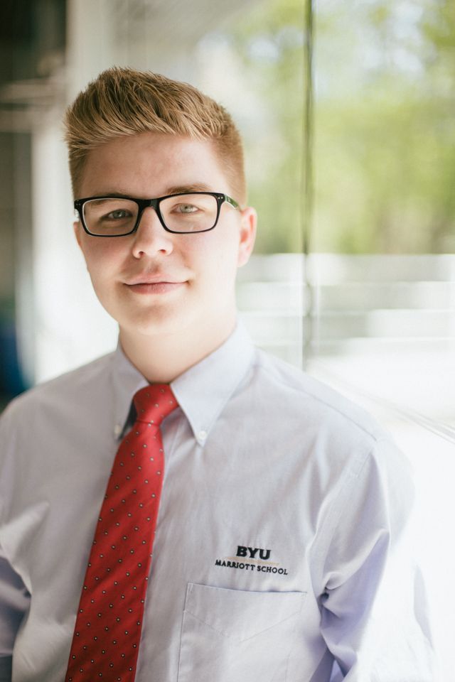A person wearing a light blue dress shirt, red tie, and glasses with a BYU Marriott School logo standing near a window.