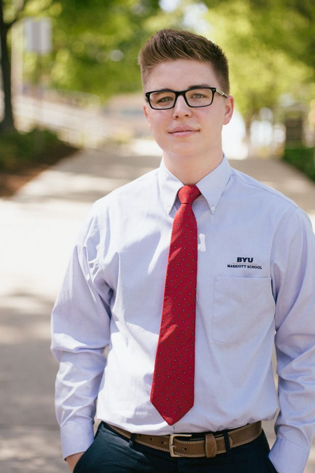 A person wearing a light blue dress shirt, red tie, and glasses stands outdoors on a walkway with blurred trees behind.