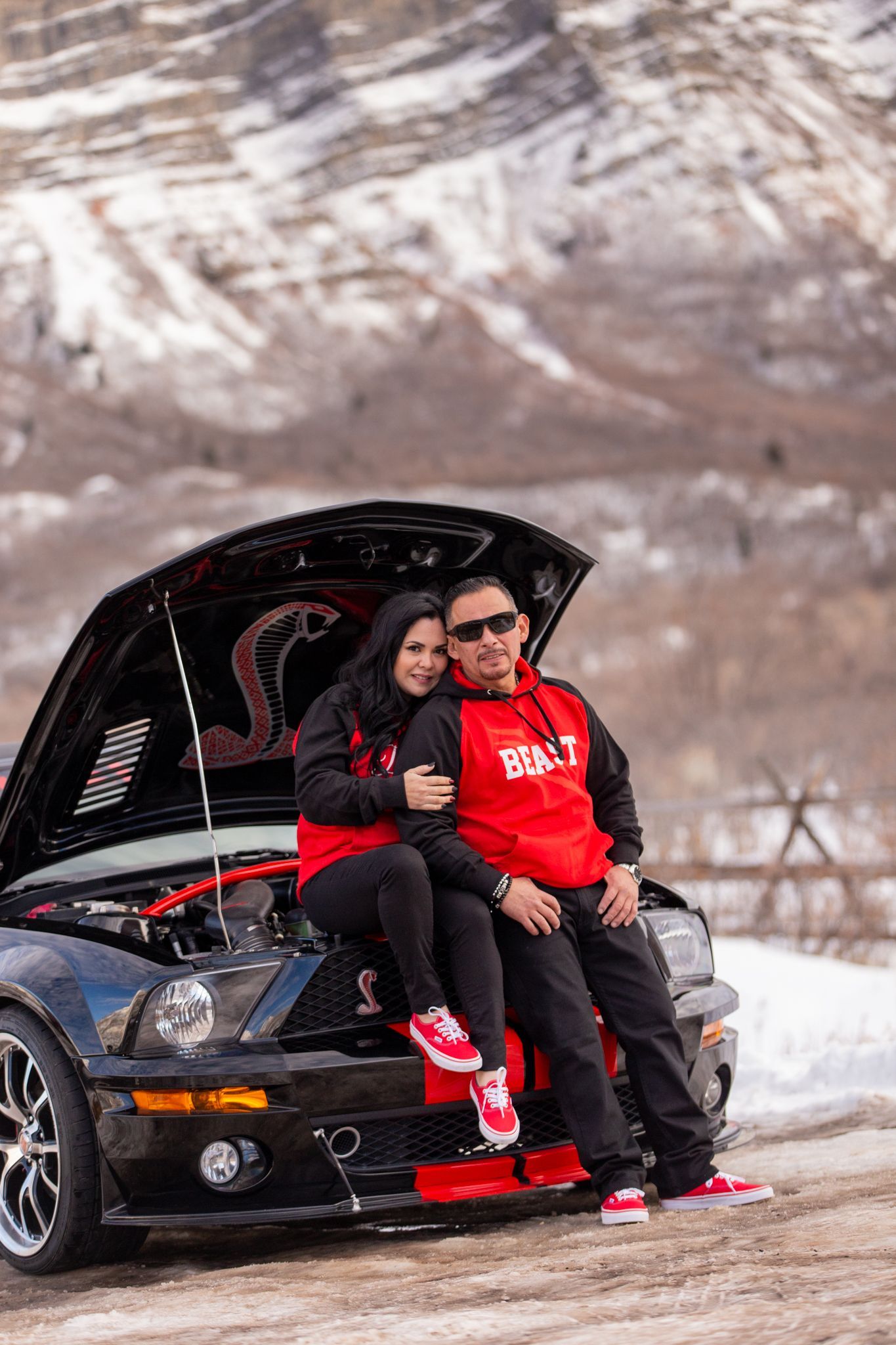 A couple poses on the front bumper of a black Ford Mustang with the hood raised, set against a snowy mountain backdrop.