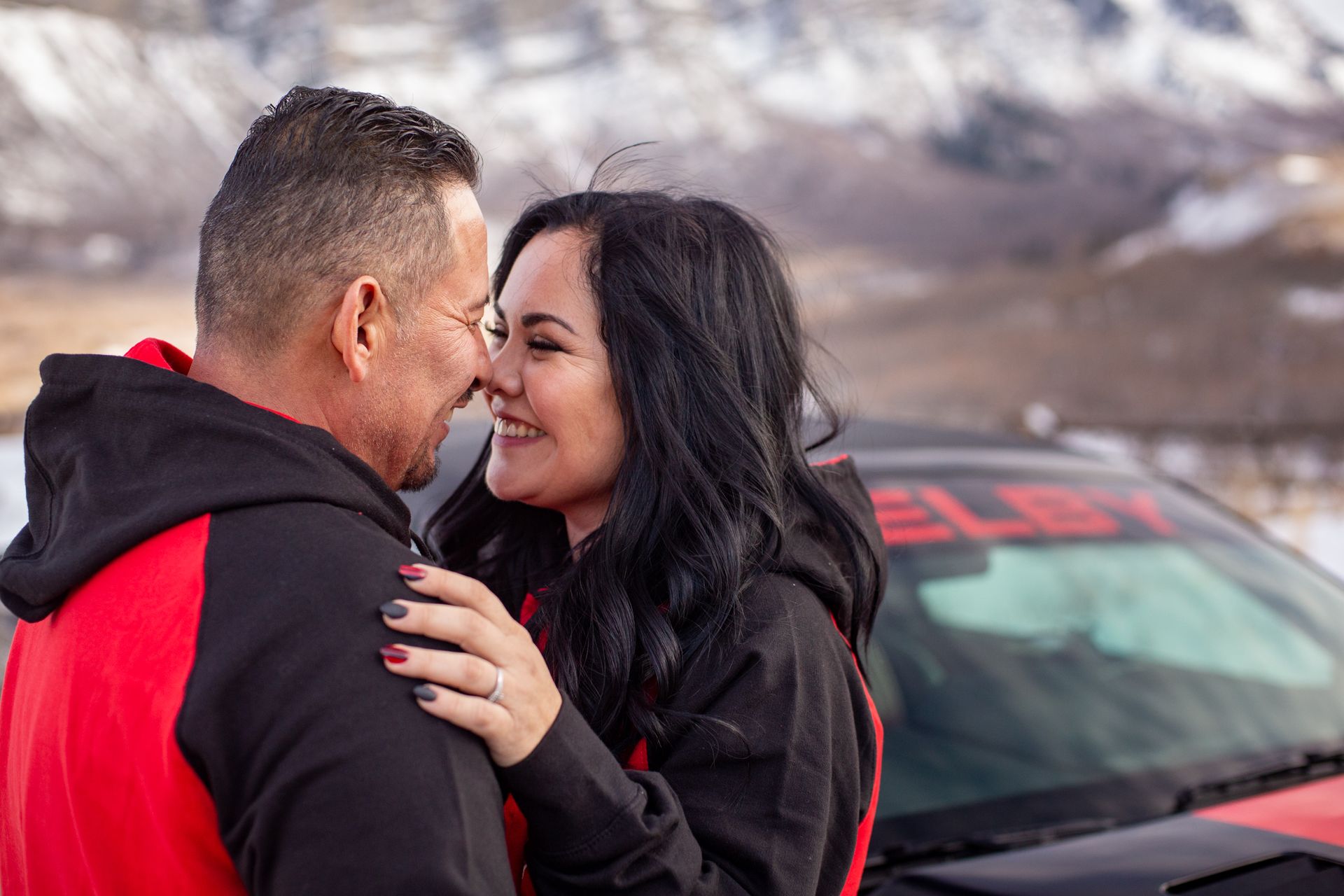 A couple smiling at each other, nose-to-nose, standing in front of a black Shelby car with snowy mountains in the distance.