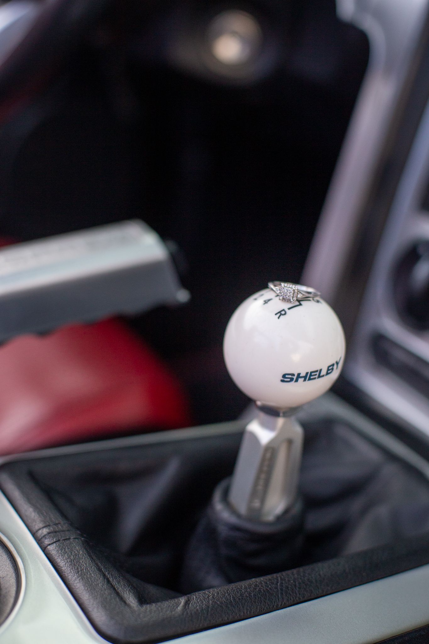 Close-up of a white Shelby-branded gear shifter knob inside a car with a red seat visible in the background.