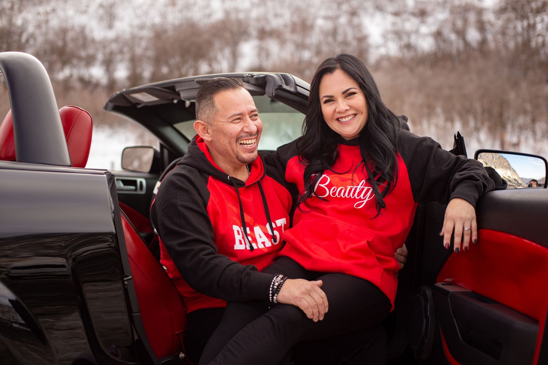 A smiling couple wearing matching red and black hoodies sits together in a convertible car on a snowy day.