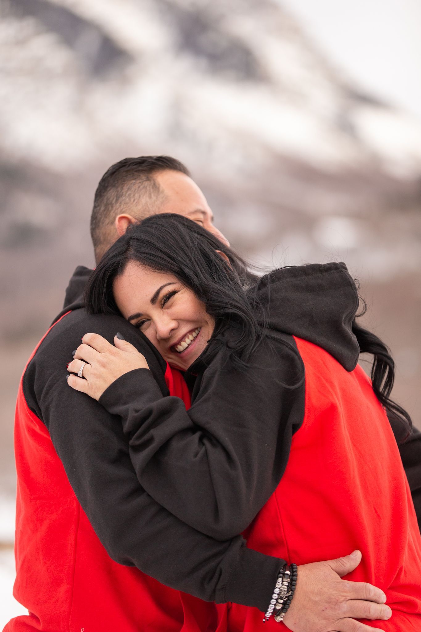 Two people embrace warmly while wearing matching red and black hoodies outdoors with a snowy, blurred mountain background.