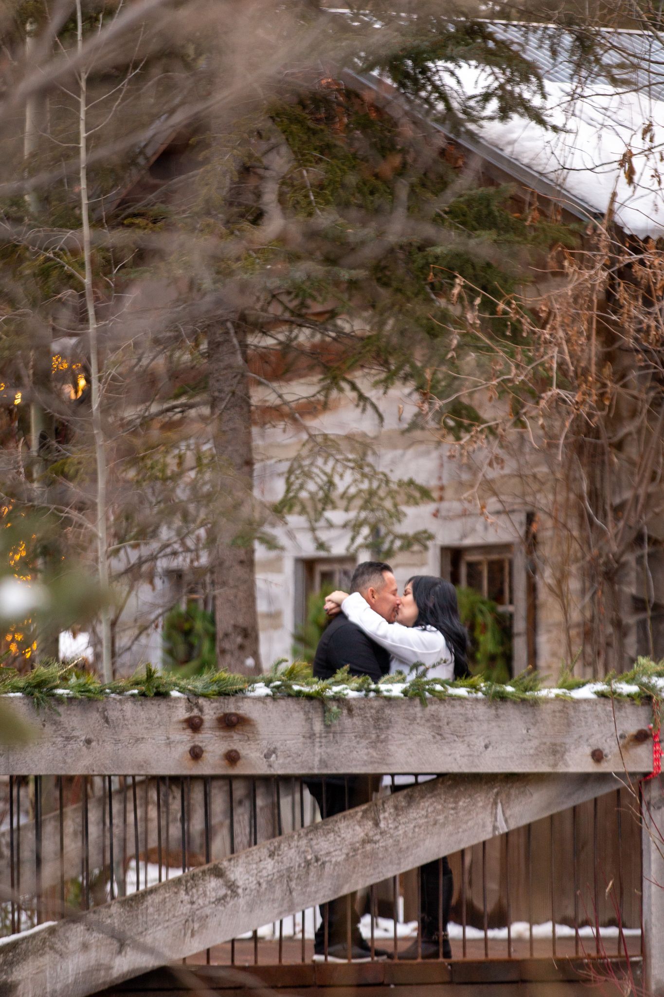 A couple kisses on a wooden bridge in front of a stone building during a snowy winter day.