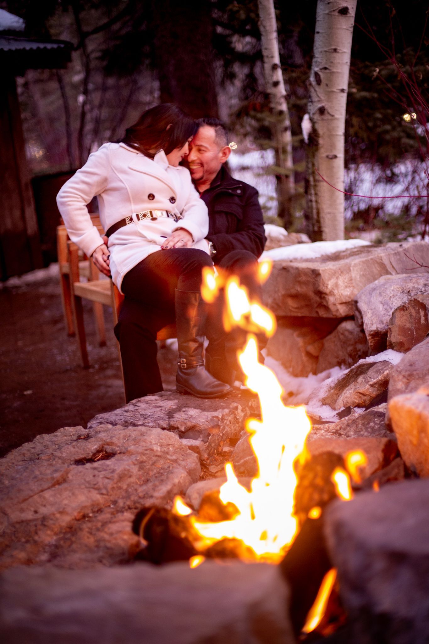 A couple sits close together on rocks by a fire pit in a snowy outdoor setting, looking at each other affectionately.