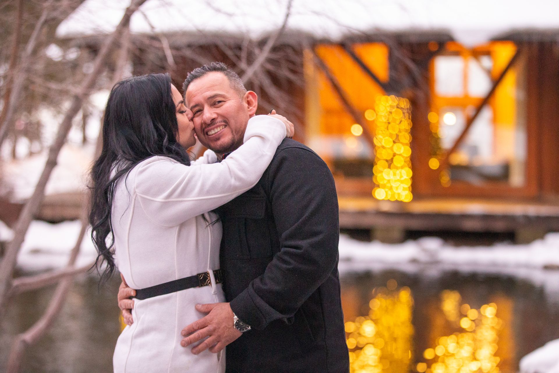 A couple embraces near a pond, with a person kissing their partner on the cheek in front of a glowing, snowy cabin.