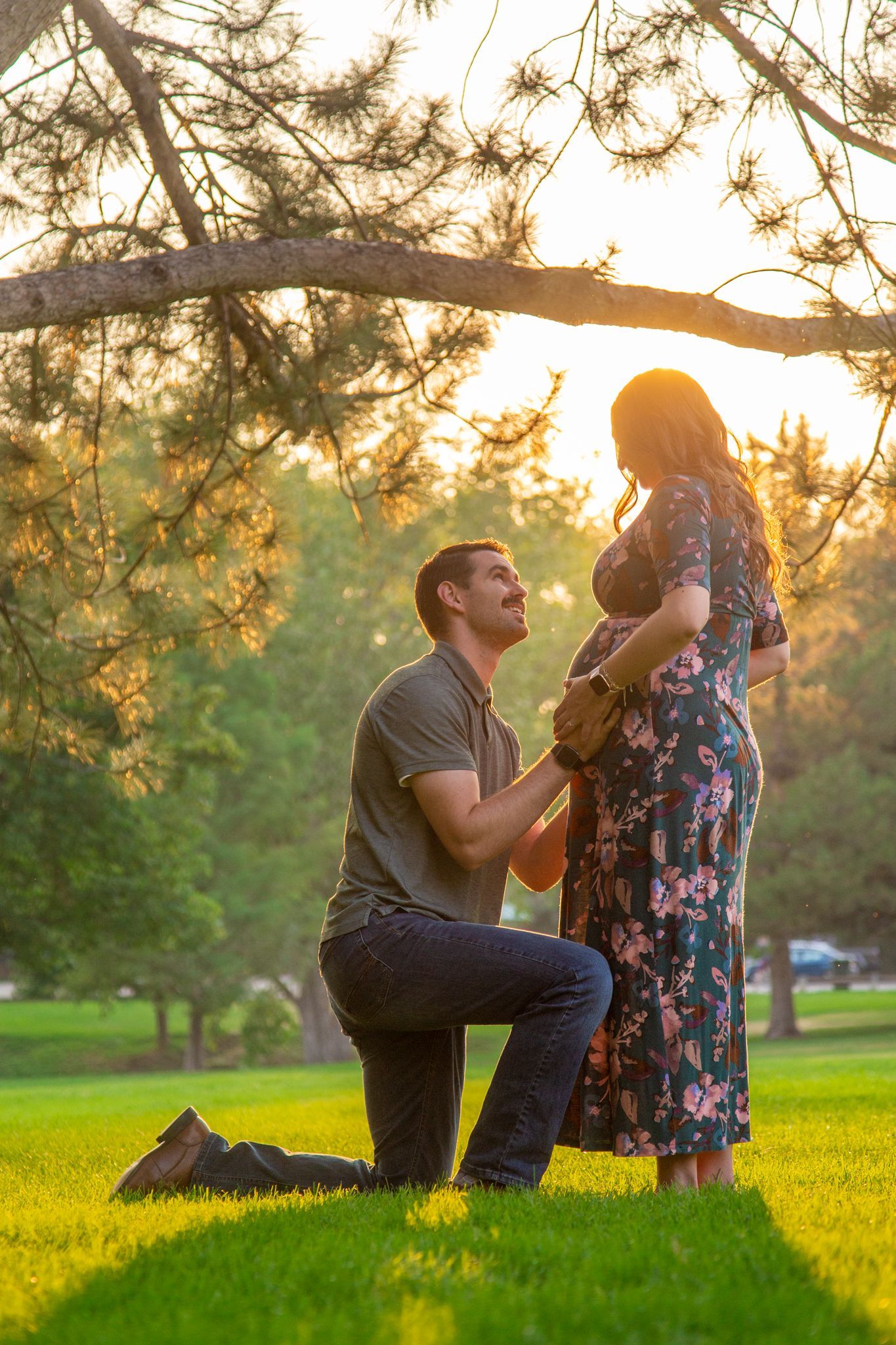 A man kneels before a pregnant person in a floral dress during a sunset in a park.