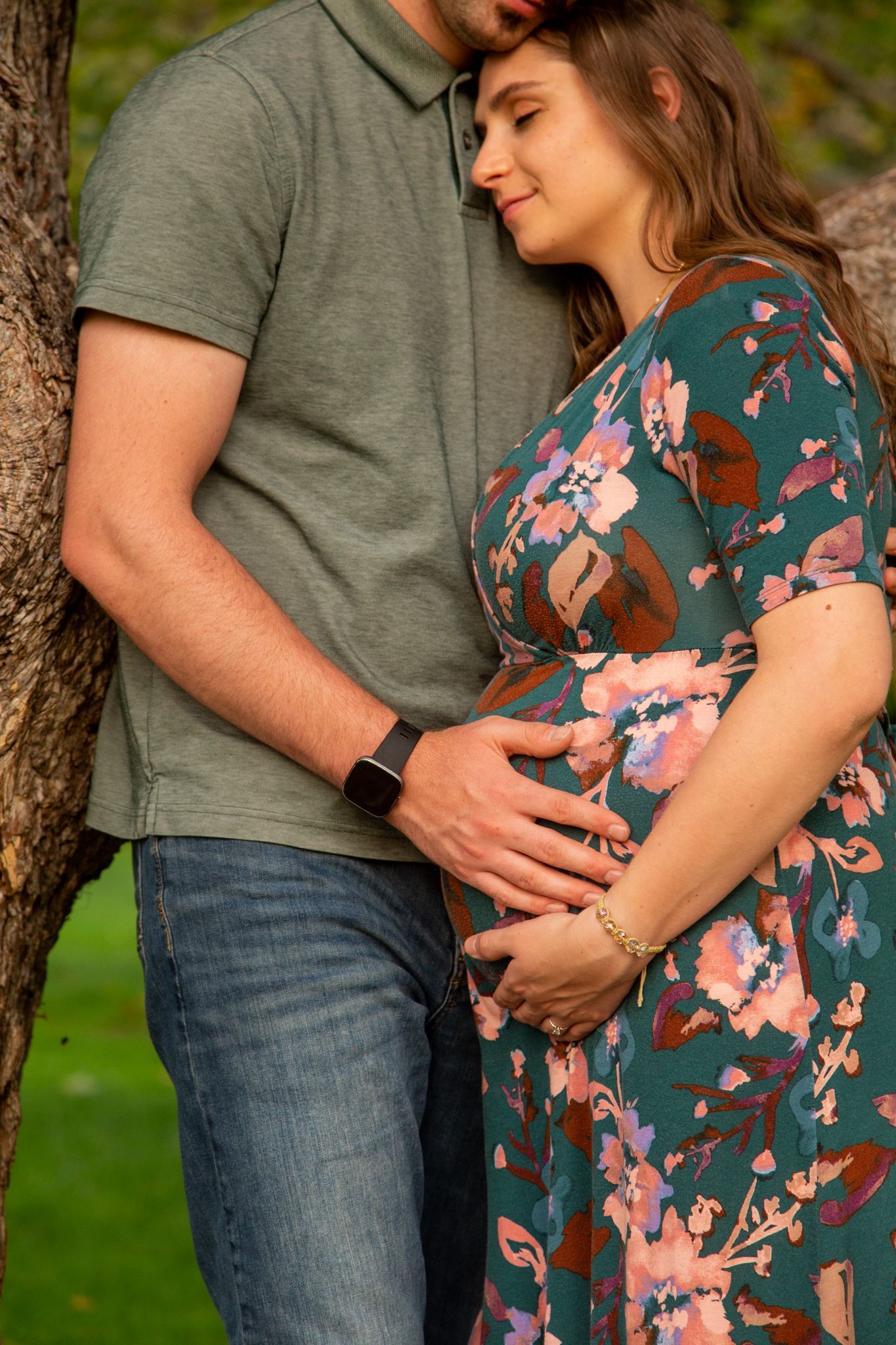 A pregnant person in a floral dress and a partner in a green polo shirt embrace outdoors next to a tree.