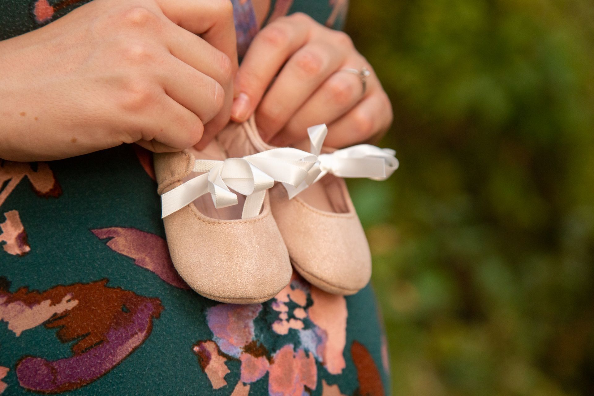 A person wears a dark floral dress and holds a pair of small, soft-pink baby shoes with white ribbon ties.