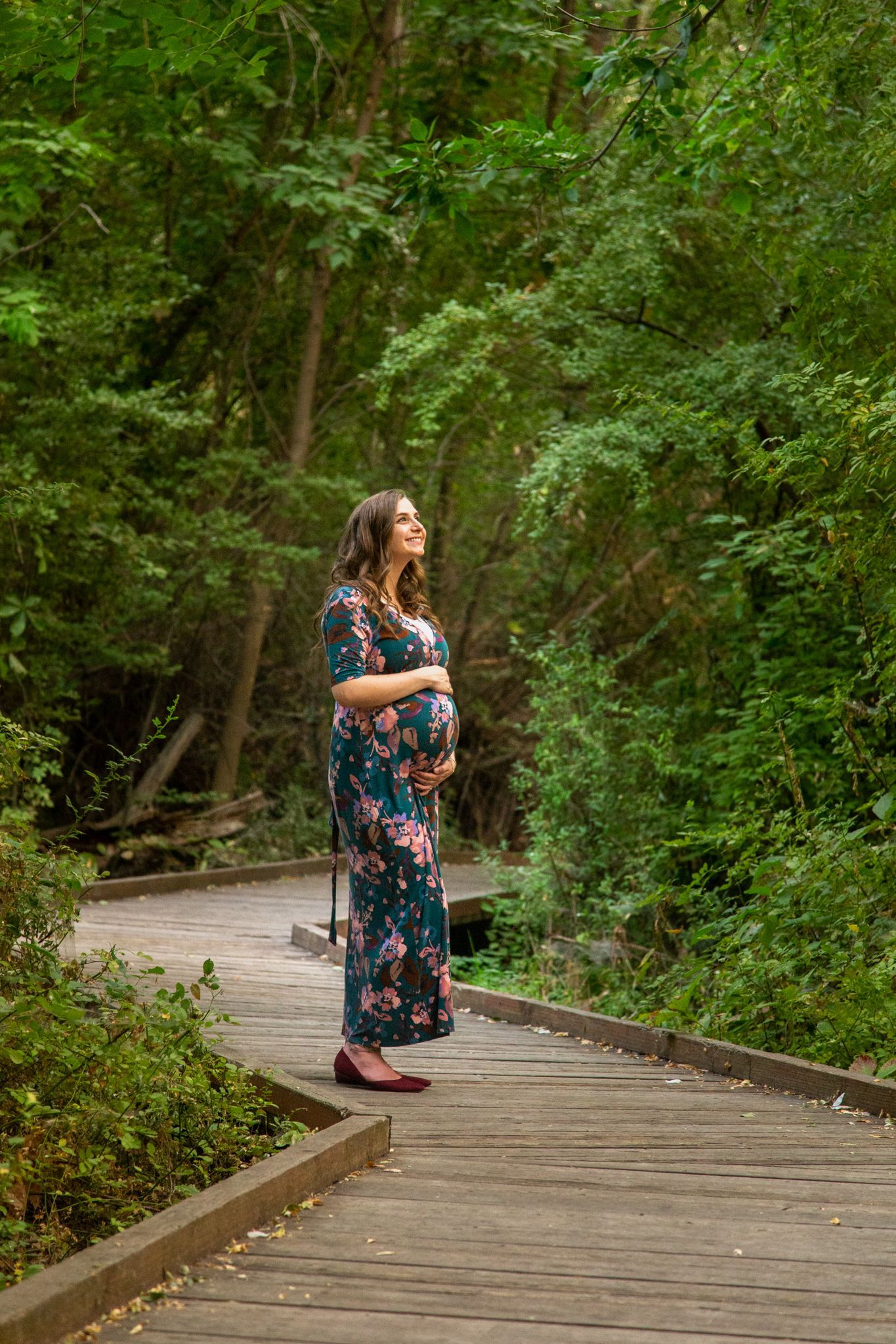 A pregnant person in a floral dress stands on a wooden boardwalk in a forest, looking upward with hands on their belly.