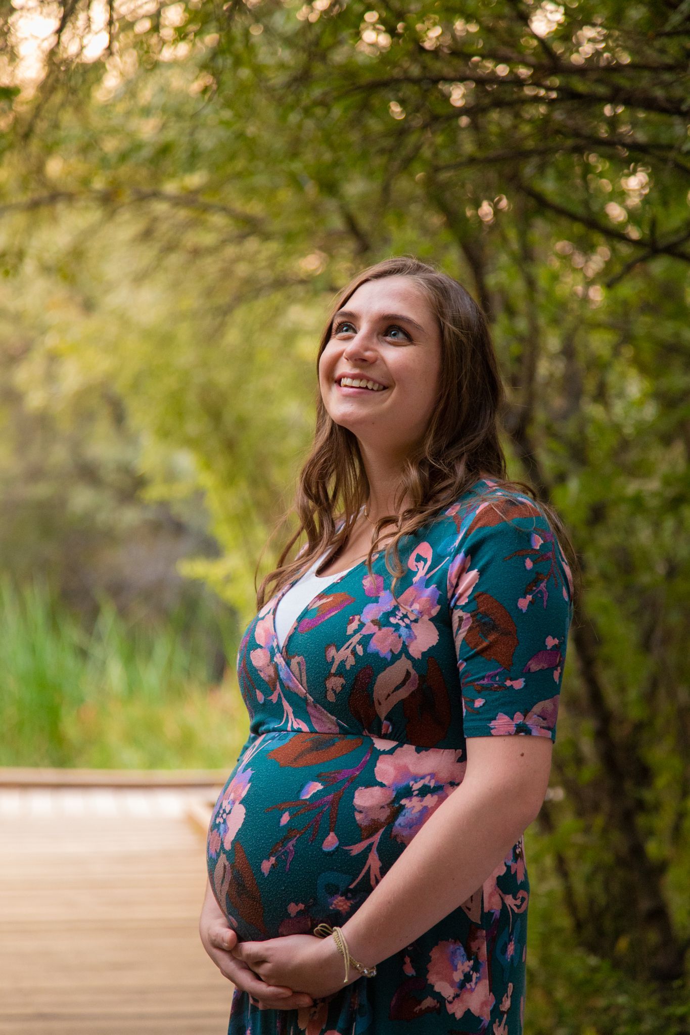 A person with long brown hair wearing a floral dress stands outdoors, cradling their baby bump and smiling upward.