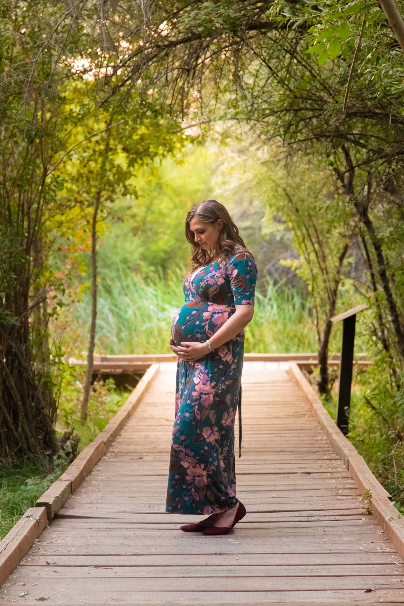 A pregnant person in a teal floral dress stands on a wooden boardwalk path surrounded by lush trees and greenery.