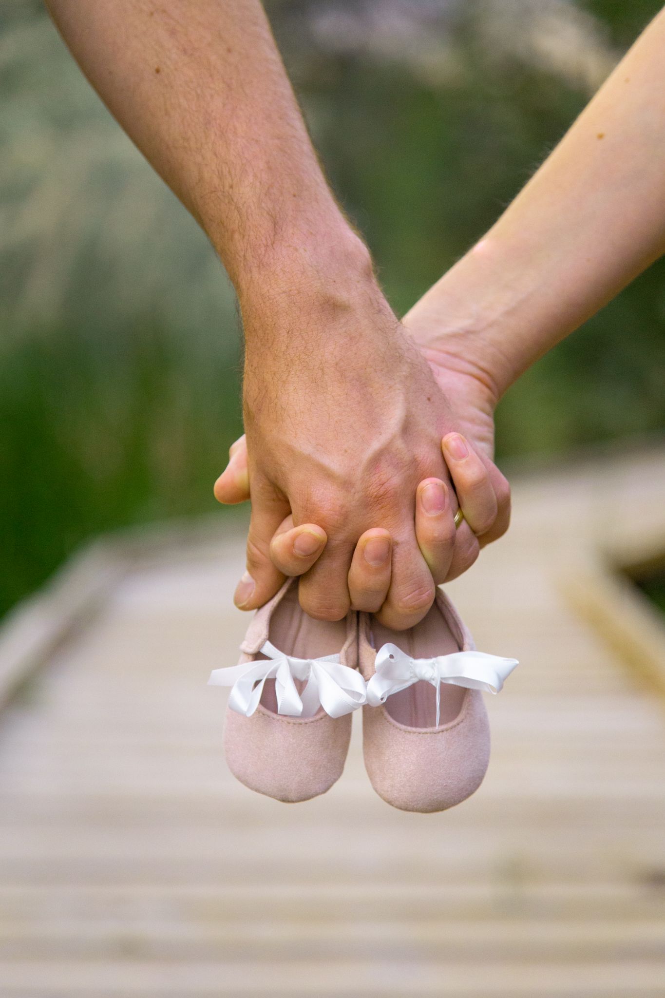Two people holding hands over a wooden bridge, dangling a pair of small, light pink baby shoes with white ribbons.