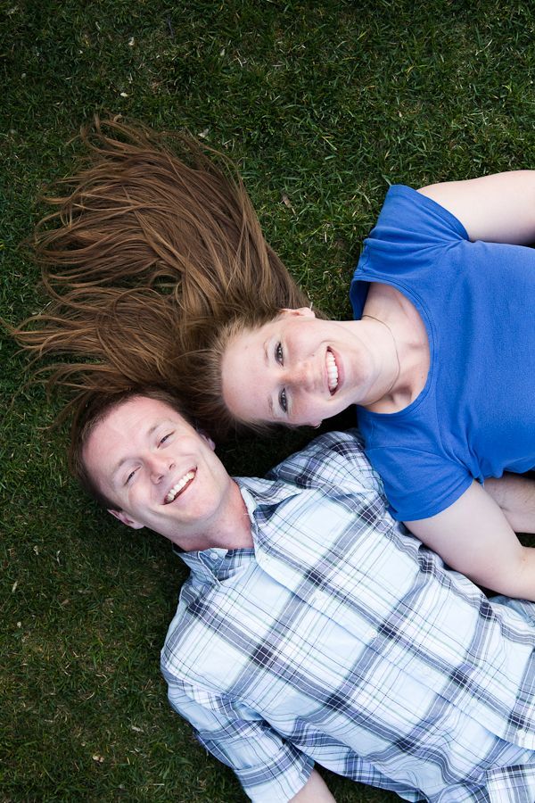 A person in a blue shirt and another in a plaid shirt lie on green grass, heads together, smiling at the camera.