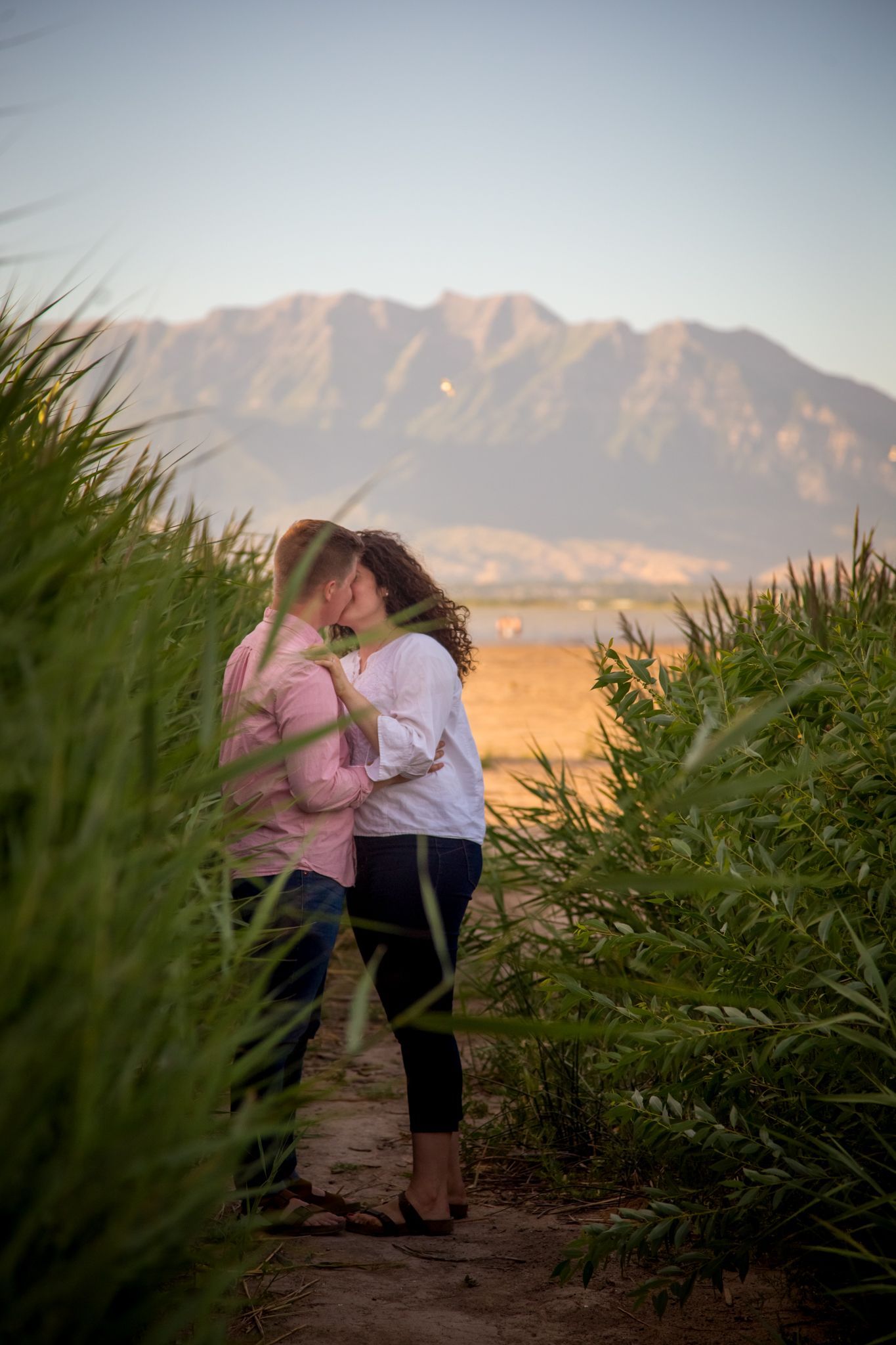 A couple stands between tall, green grasses in a natural landscape, kissing with mountains in the background.