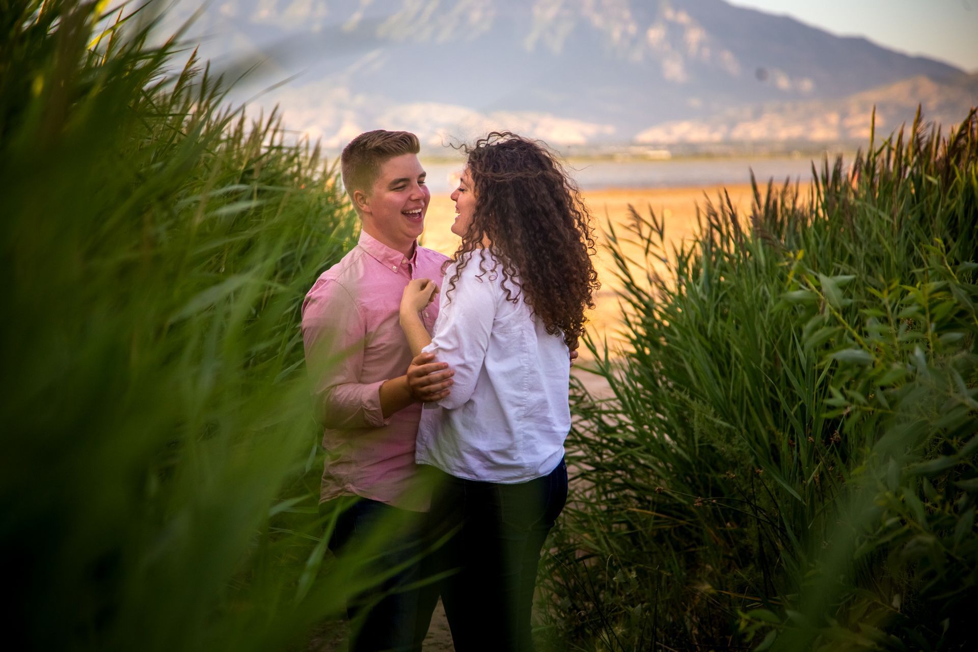 A couple laughing together while standing between tall green grass with a lake and mountains in the background.