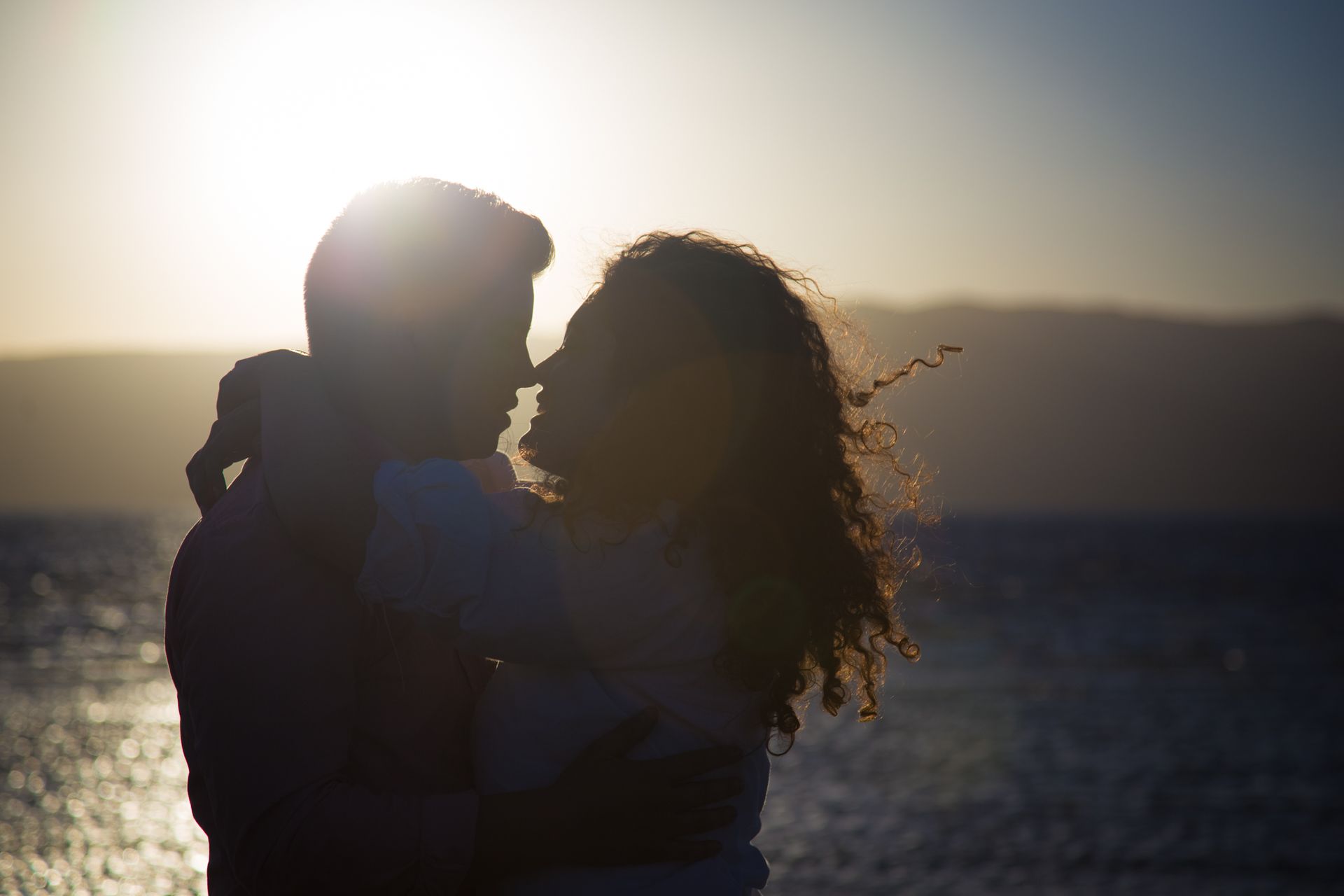 A silhouette of two people embracing and looking at each other against a bright, hazy sunset over a body of water.