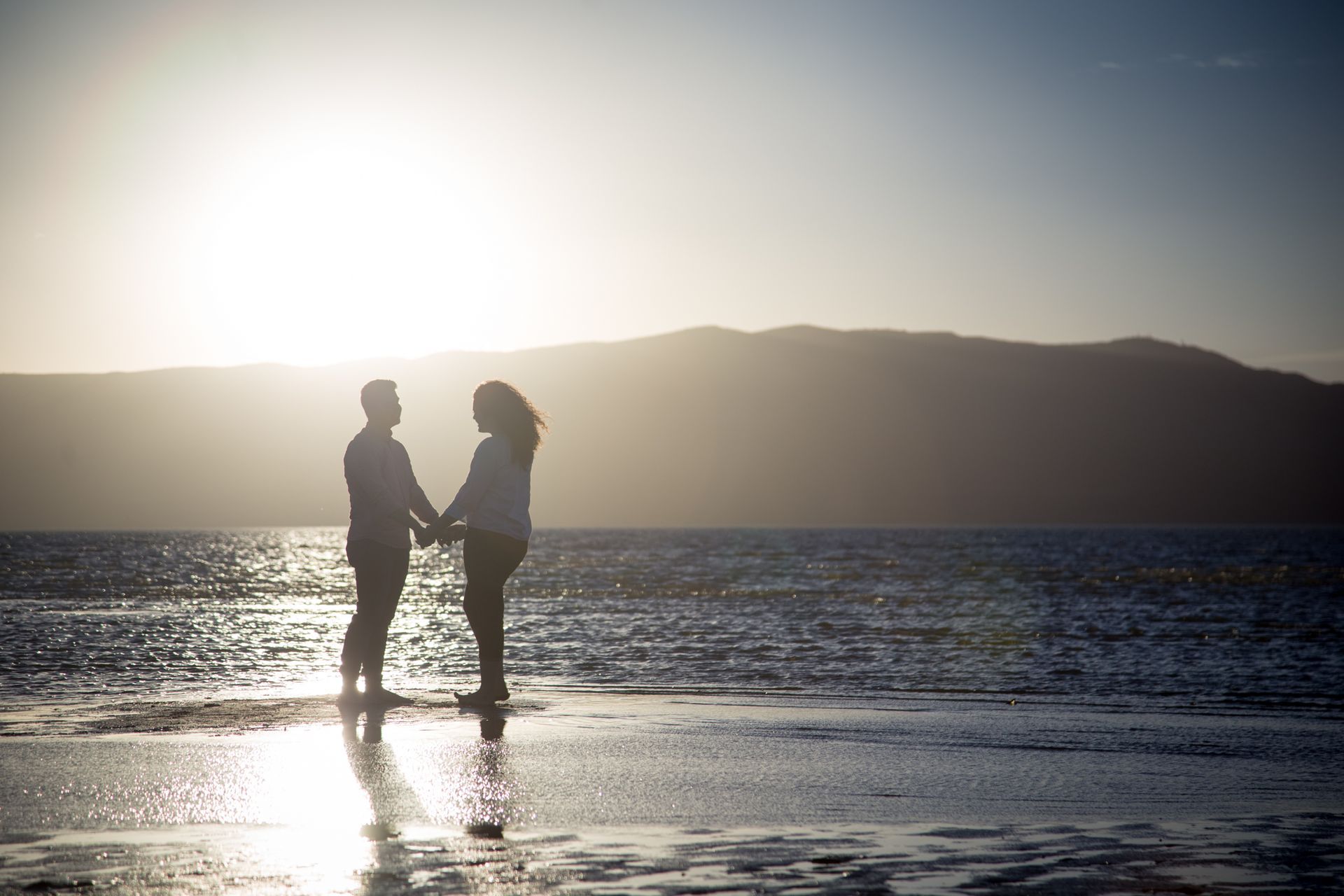 A couple stands holding hands on a beach during sunset with mountains in the background.