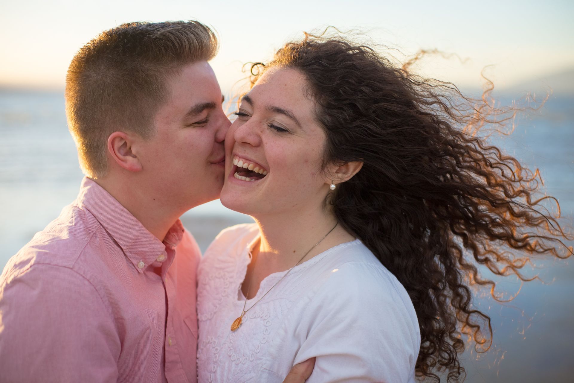 A person kisses the cheek of a laughing partner outdoors near water during golden hour.