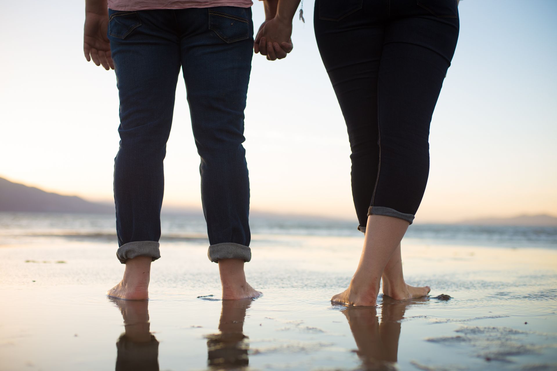 Two people holding hands while standing barefoot in shallow water on a beach at sunset.