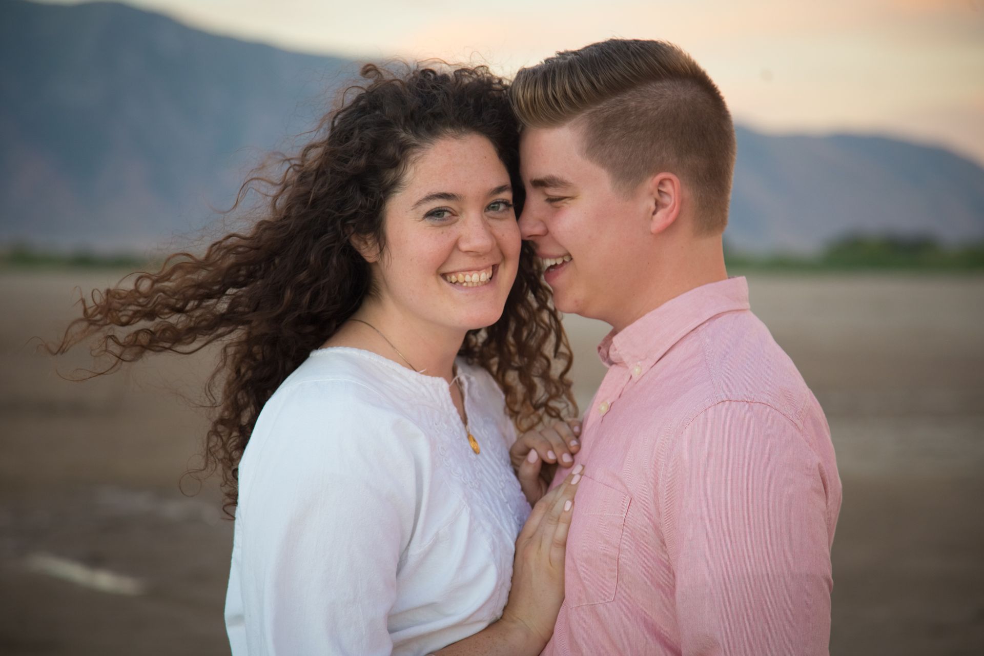 A smiling couple stands outdoors with mountains in the background, one person’s hair blowing in the wind.